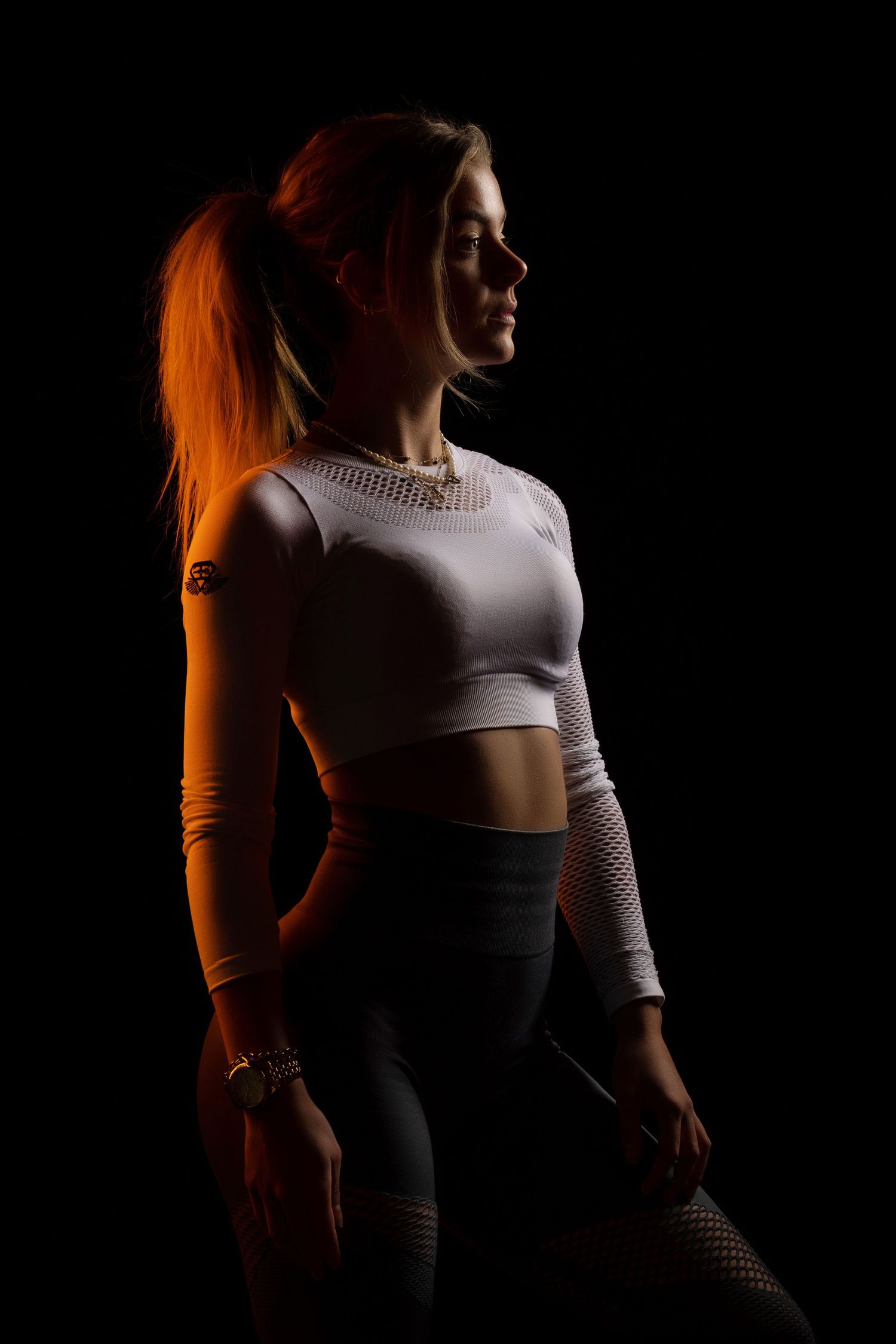 Athletic figure in white crop top and black leggings posing dramatically against dark background with orange lighting.