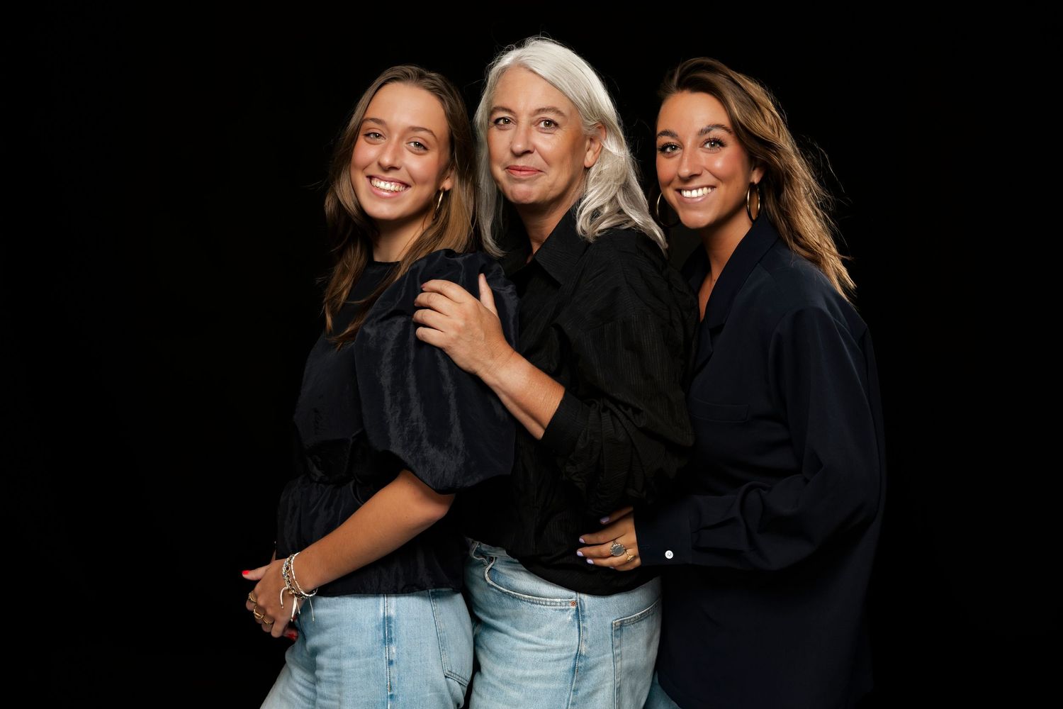 Three women wearing casual denim and dark tops pose together against a black background in studio lighting.