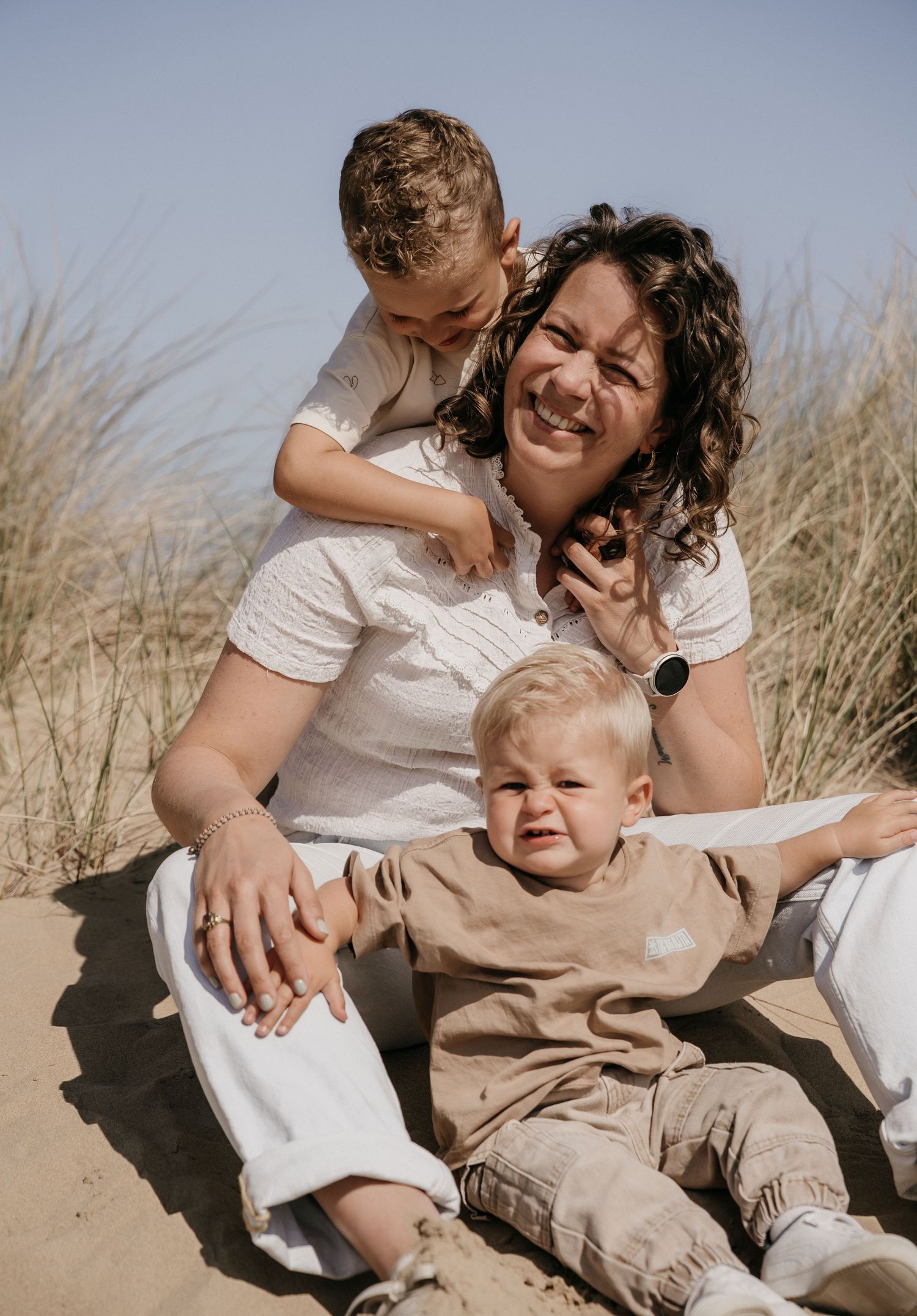A smiling woman sits on sandy dunes with two young boys, one on her back and one in her lap, surrounded by beach grass.