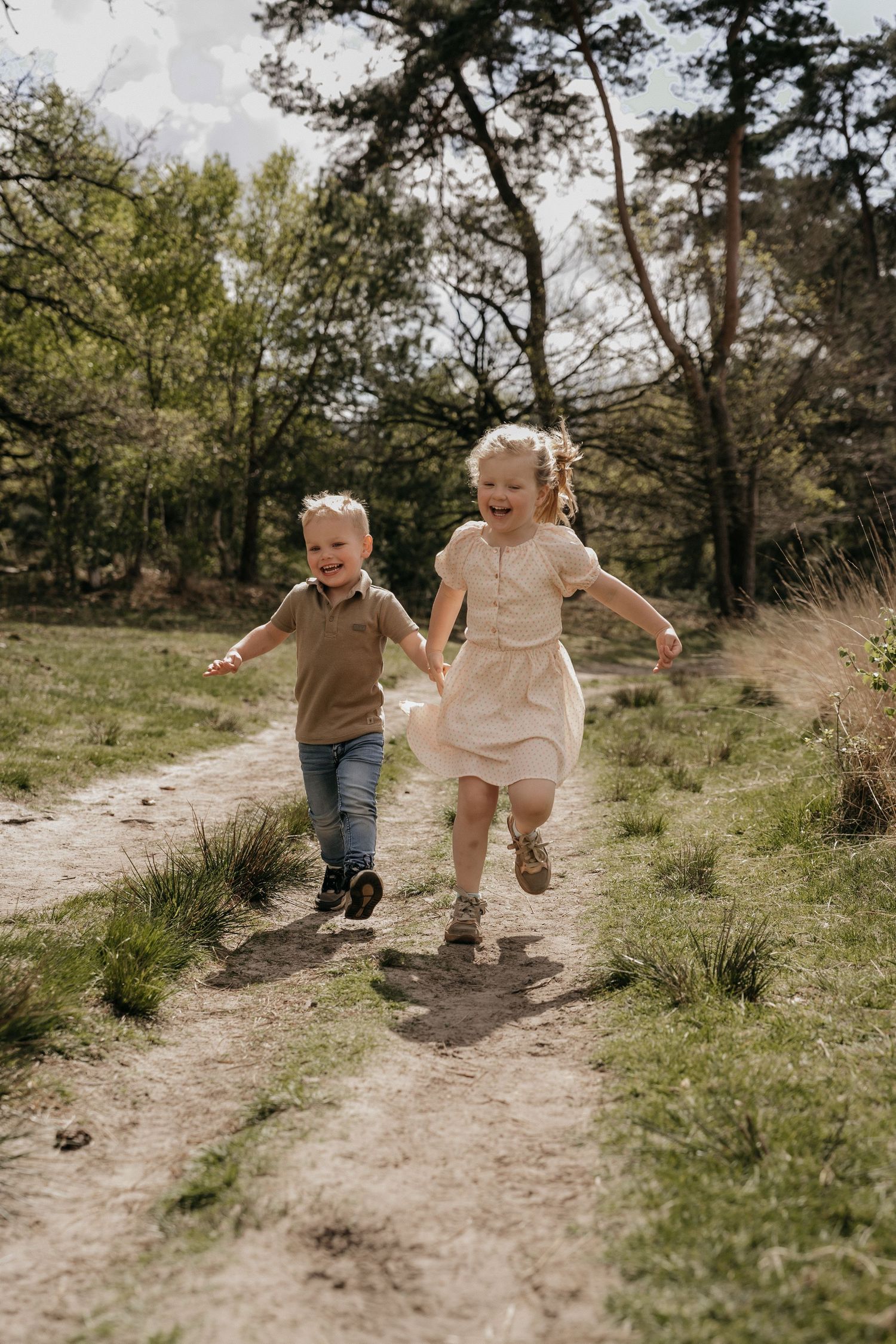 Two young children laughing and running hand in hand along a sandy forest path on a sunny day.