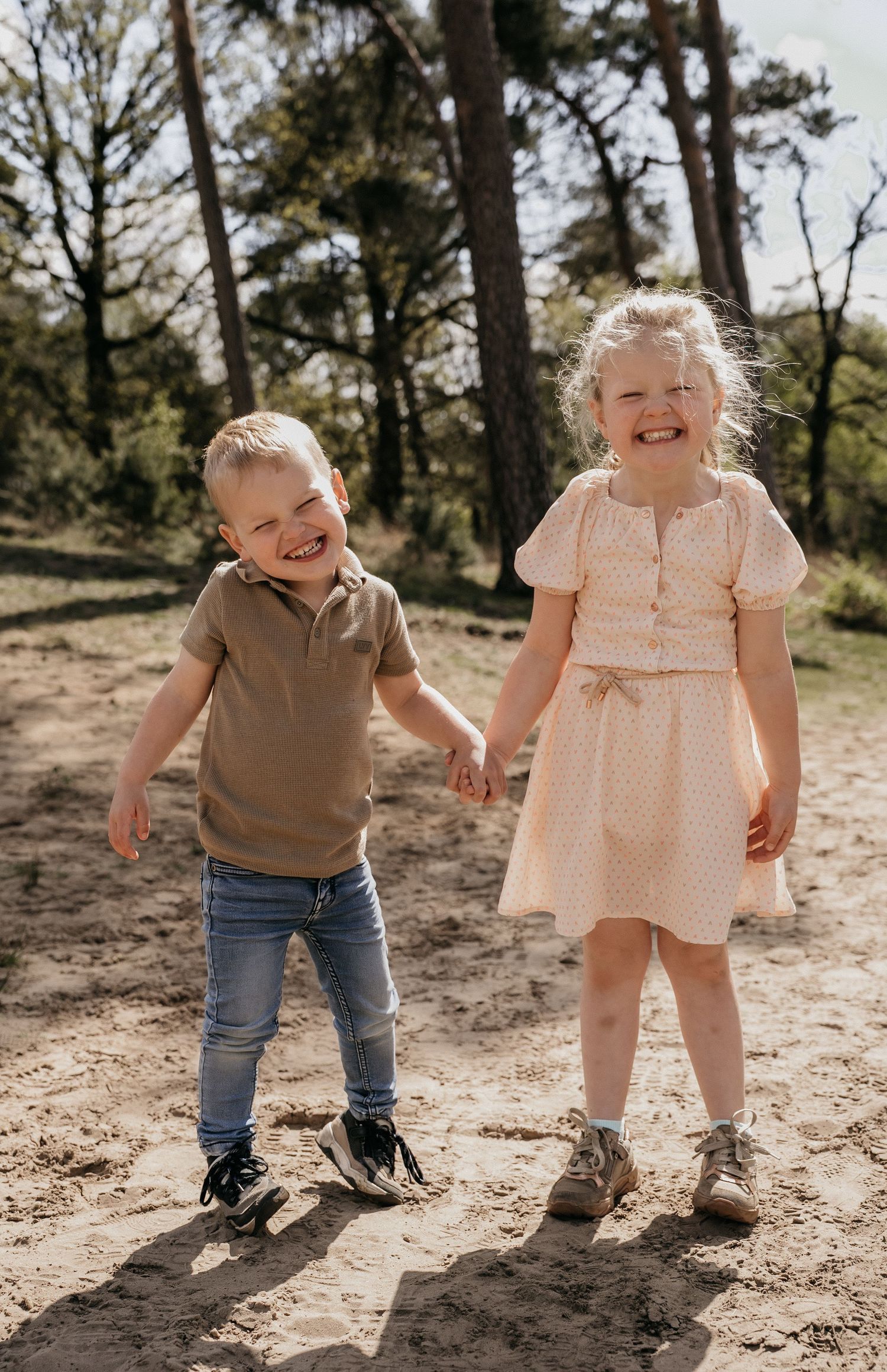 Two happy children holding hands and laughing outdoors on sandy ground surrounded by pine trees on a sunny day.