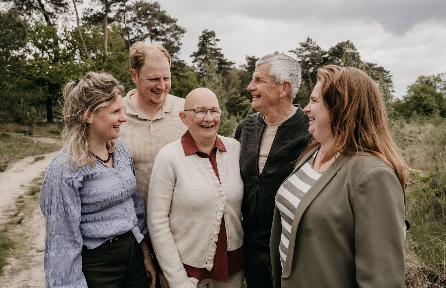 Five adults smiling and laughing together outdoors on a sandy path surrounded by trees.