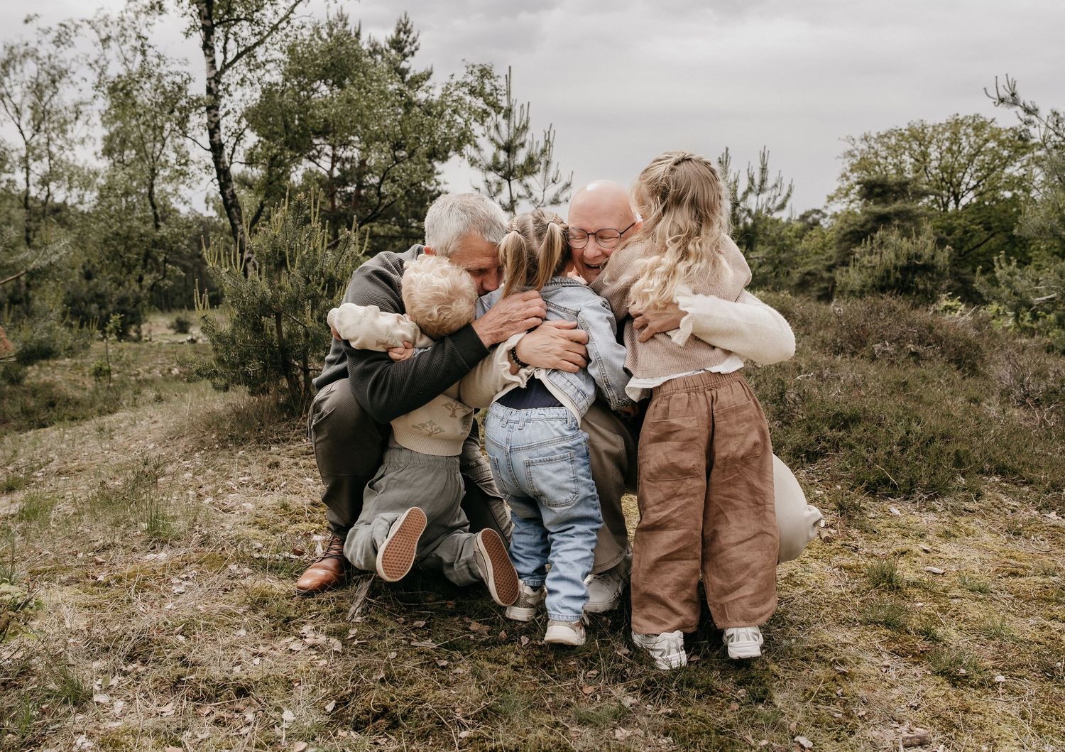 Two grandparents hugging and kissing three young grandchildren outdoors in a natural woodland setting.