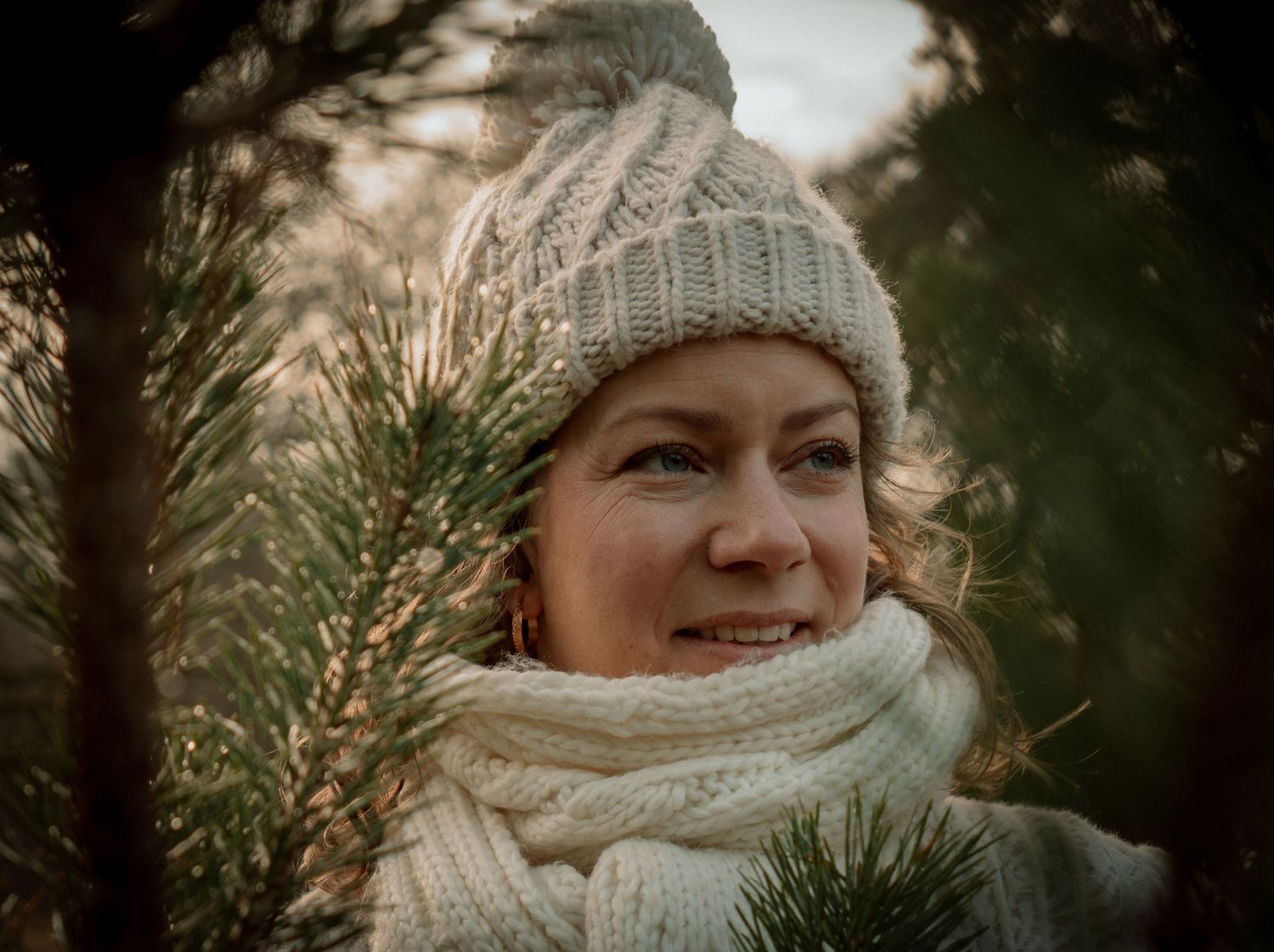 A winter scene with pine branches and a person wearing a cozy beige knit hat and scarf in natural light.