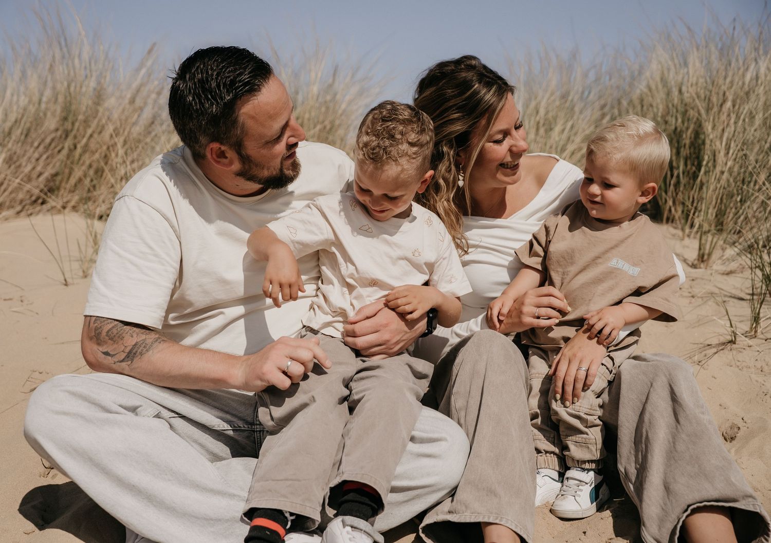Happy family of four sitting together on sandy beach dunes, parents smiling with two young children in neutral tones.