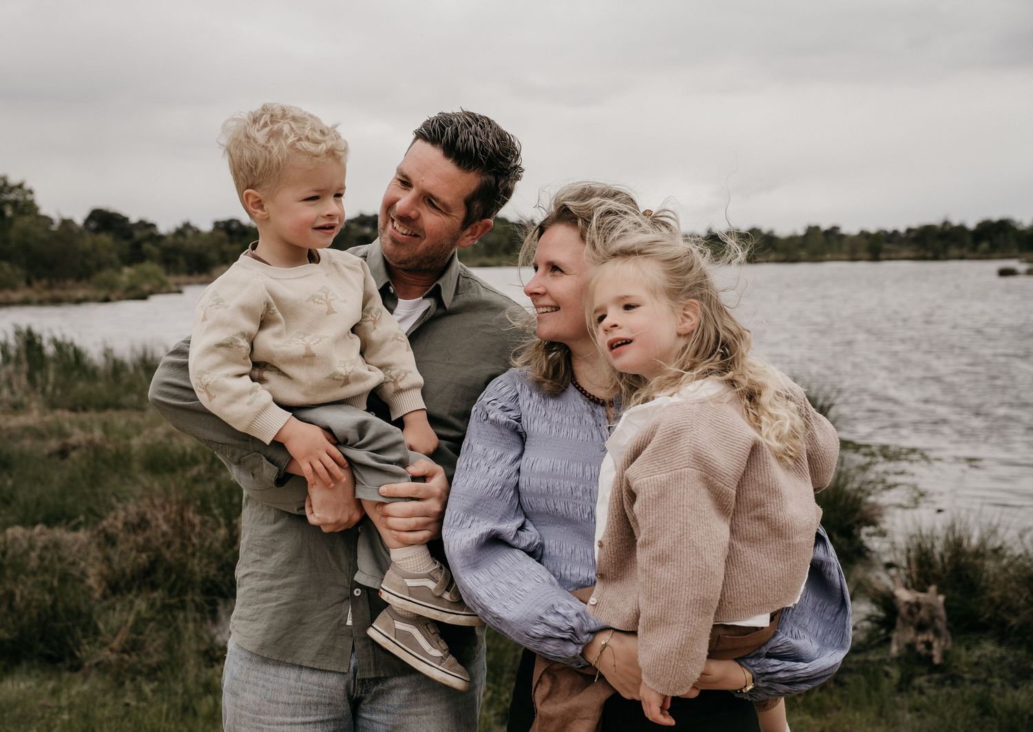 Happy family of four smiling outdoors near a lake, parents holding young children on a cloudy day.