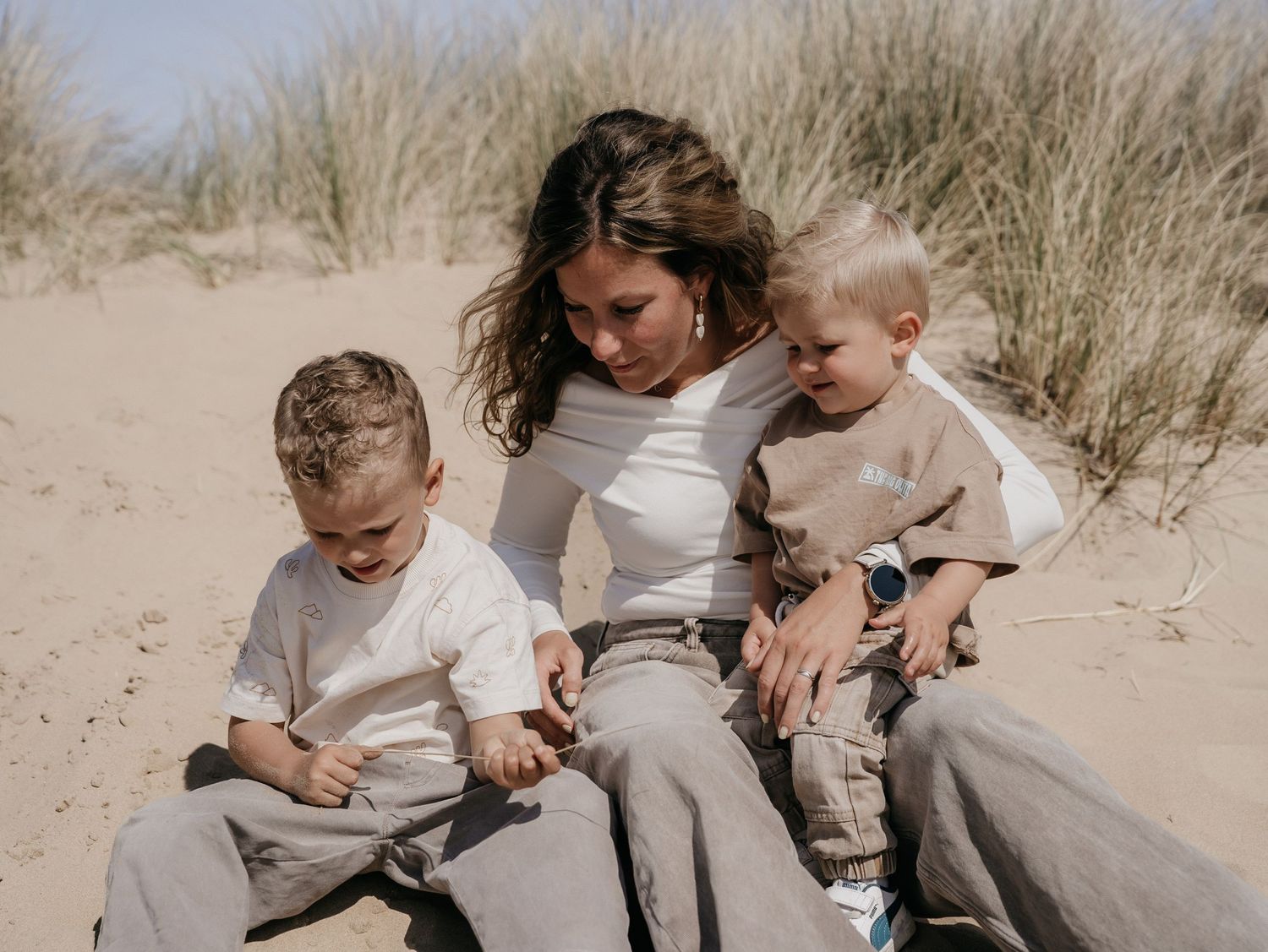 A young mother sits on sandy beach dunes with two toddler boys in neutral tones on a sunny day.