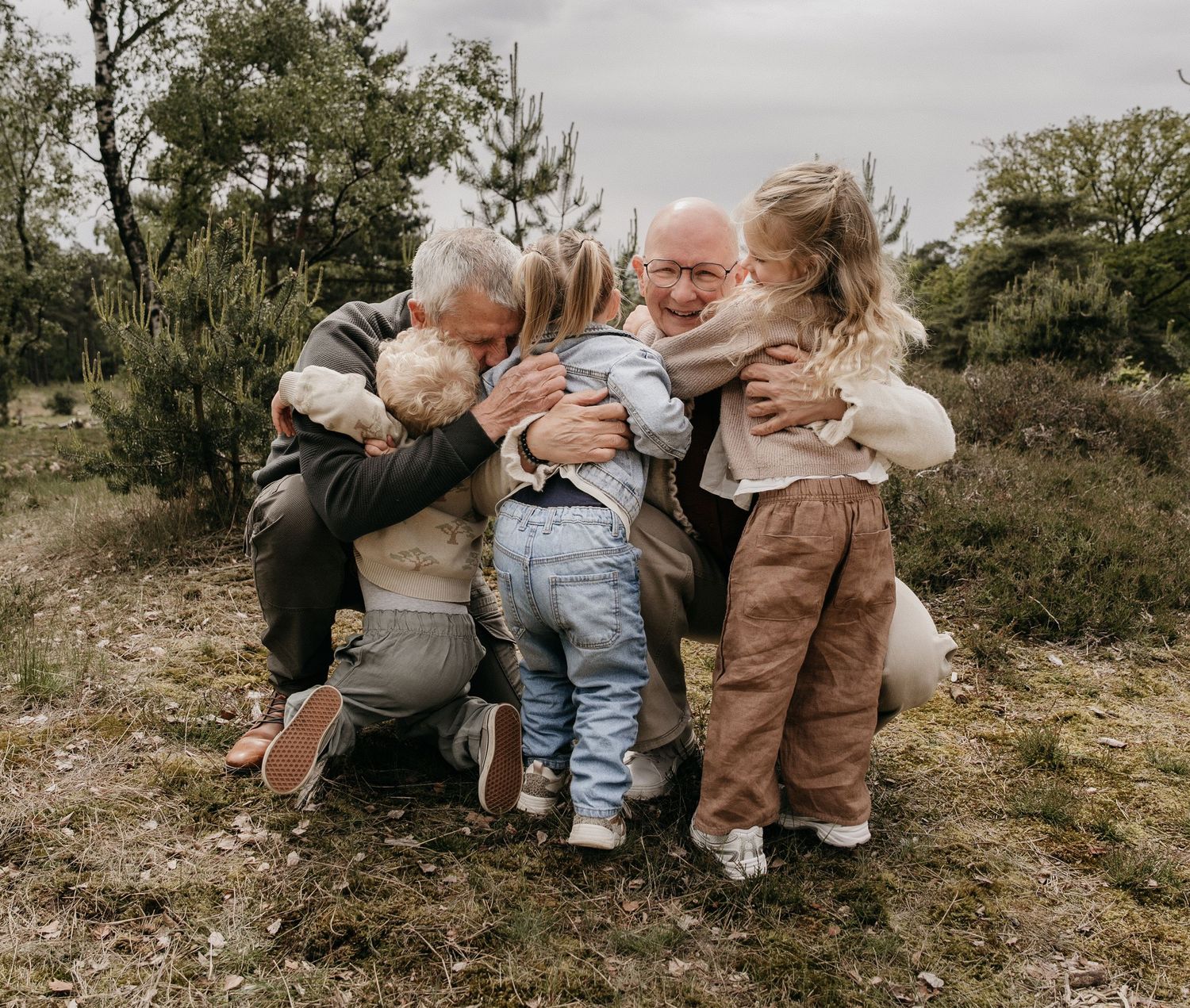 Grandparents hugging multiple young grandchildren outdoors in a natural woodland setting on an overcast day.