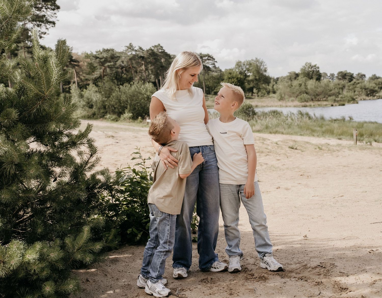 Blonde mother hugging two young boys outdoors on a sandy path surrounded by trees and a lake.