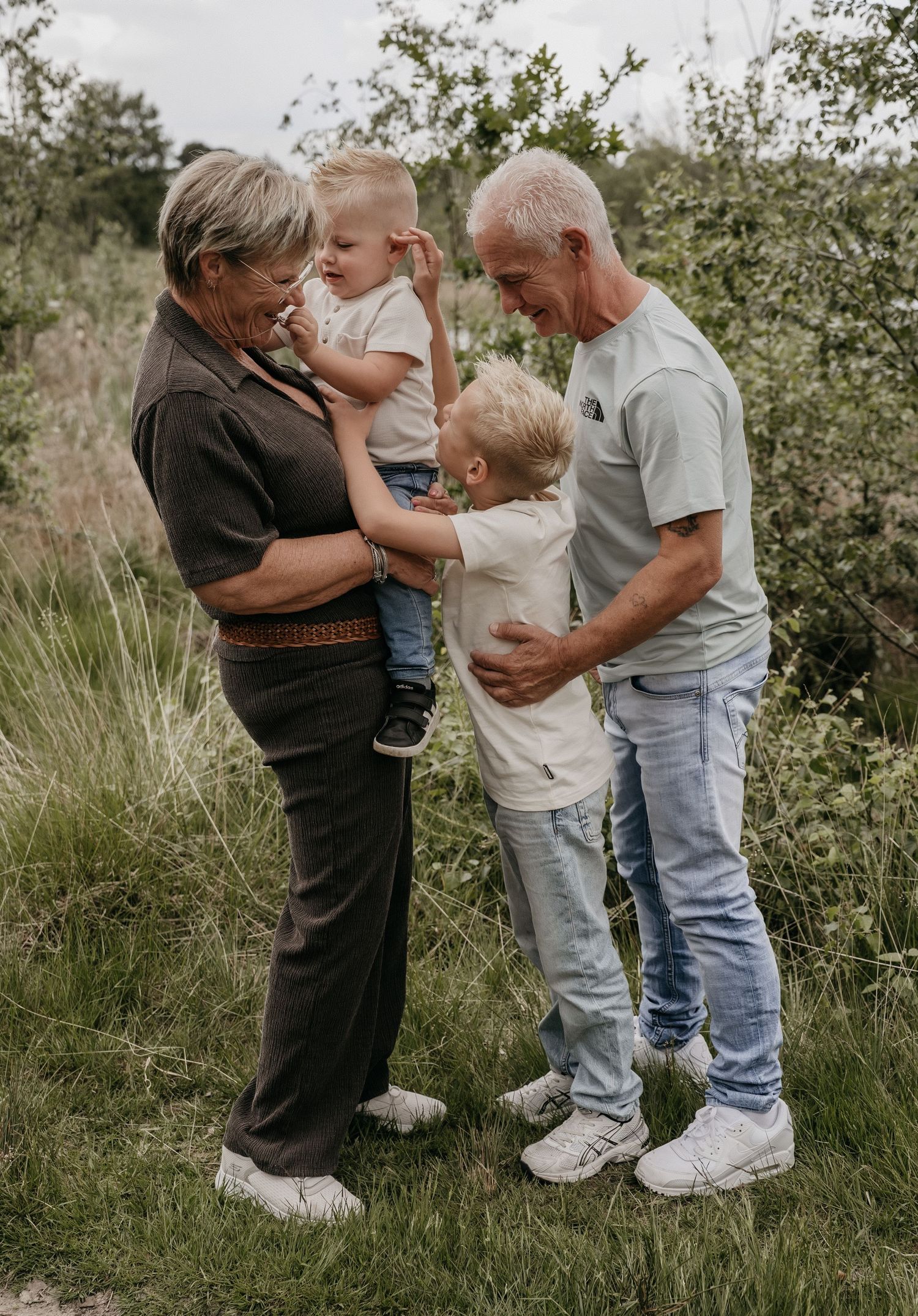 Grandparents smiling and playing with two young blonde boys outdoors in a green natural setting.