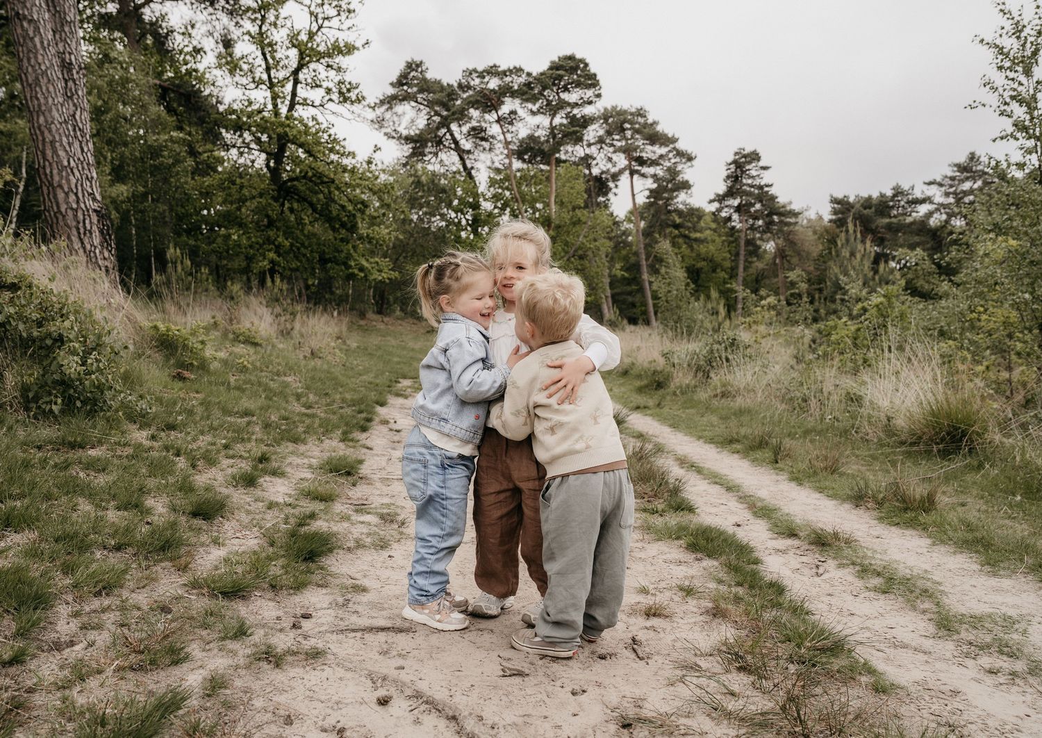 Three young children hugging each other on a sandy forest path surrounded by green trees and shrubs.