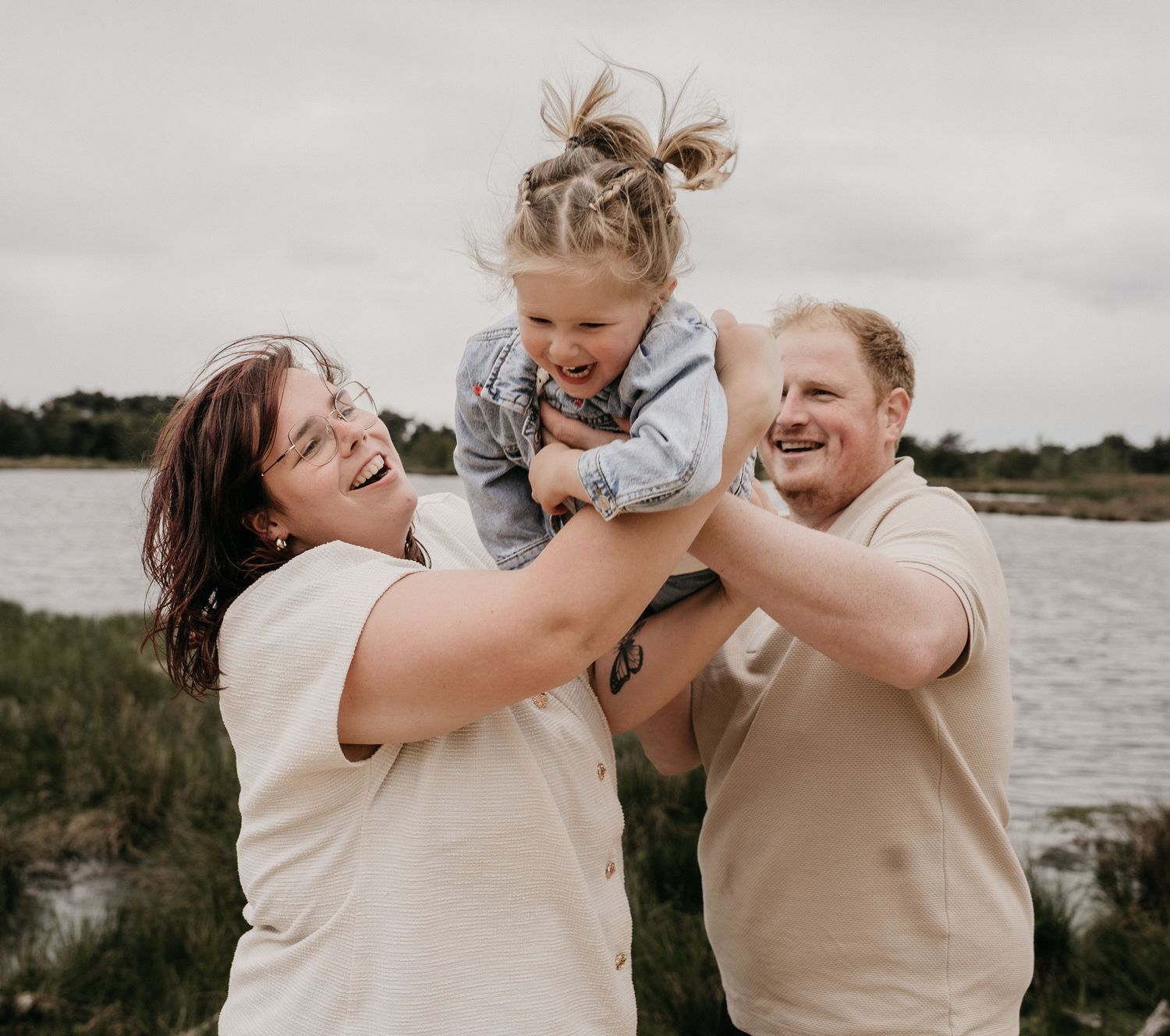 Two parents joyfully lift their laughing toddler girl outdoors near a lake on a cloudy day.