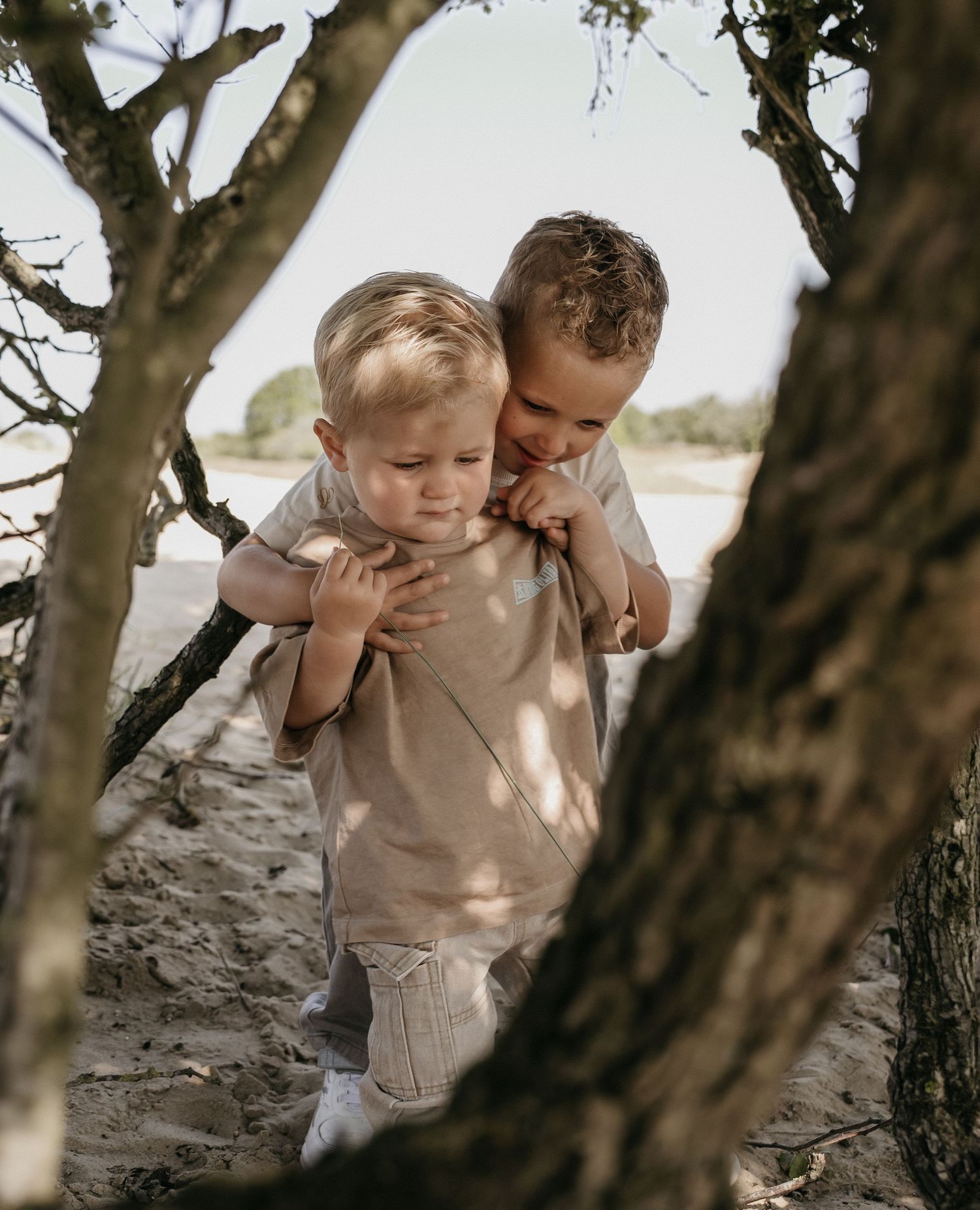 Two young boys playing together among trees on a sandy beach, older child hugging younger sibling.