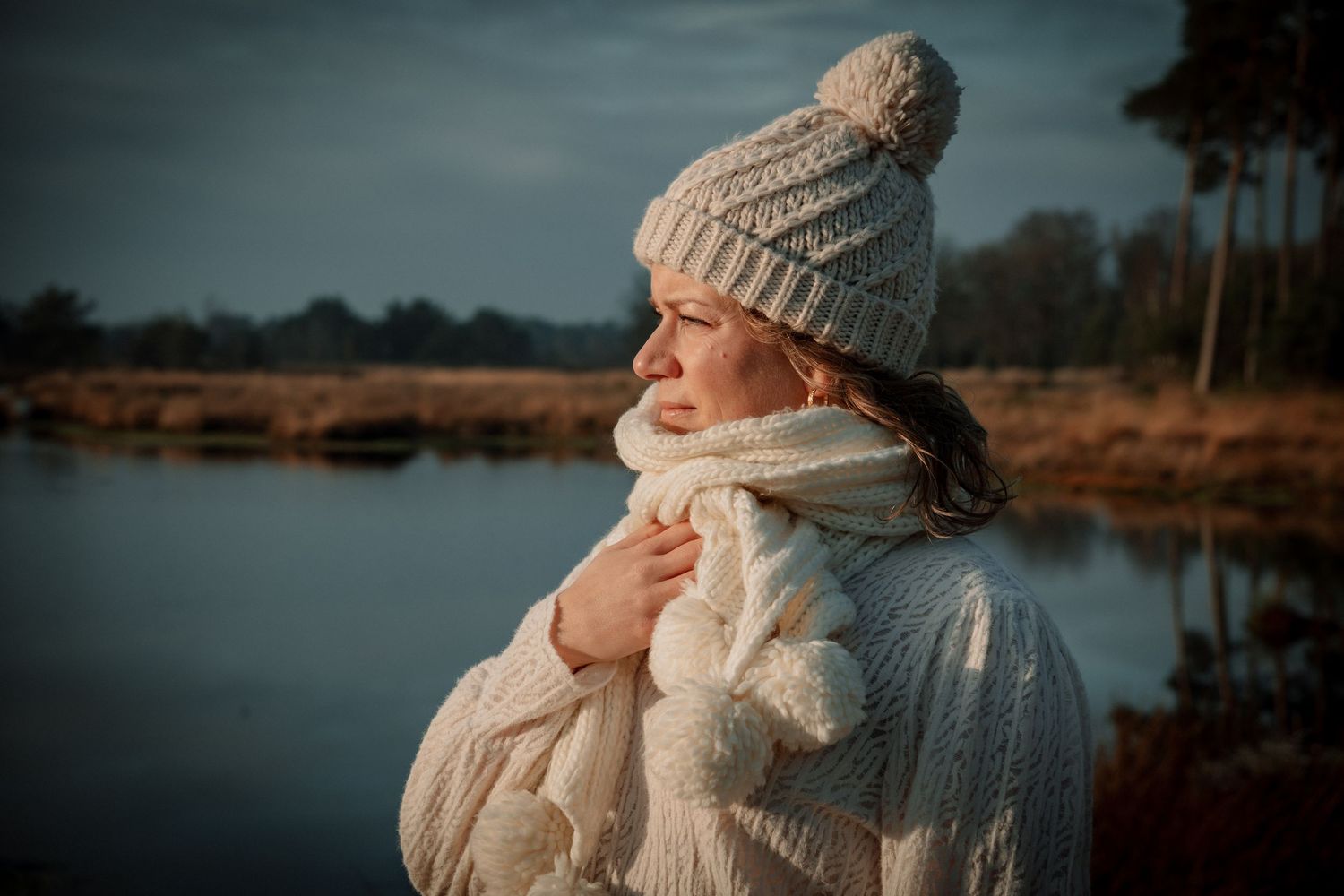 A romantic couple in warm winter clothing embraces by a serene lake during sunset.