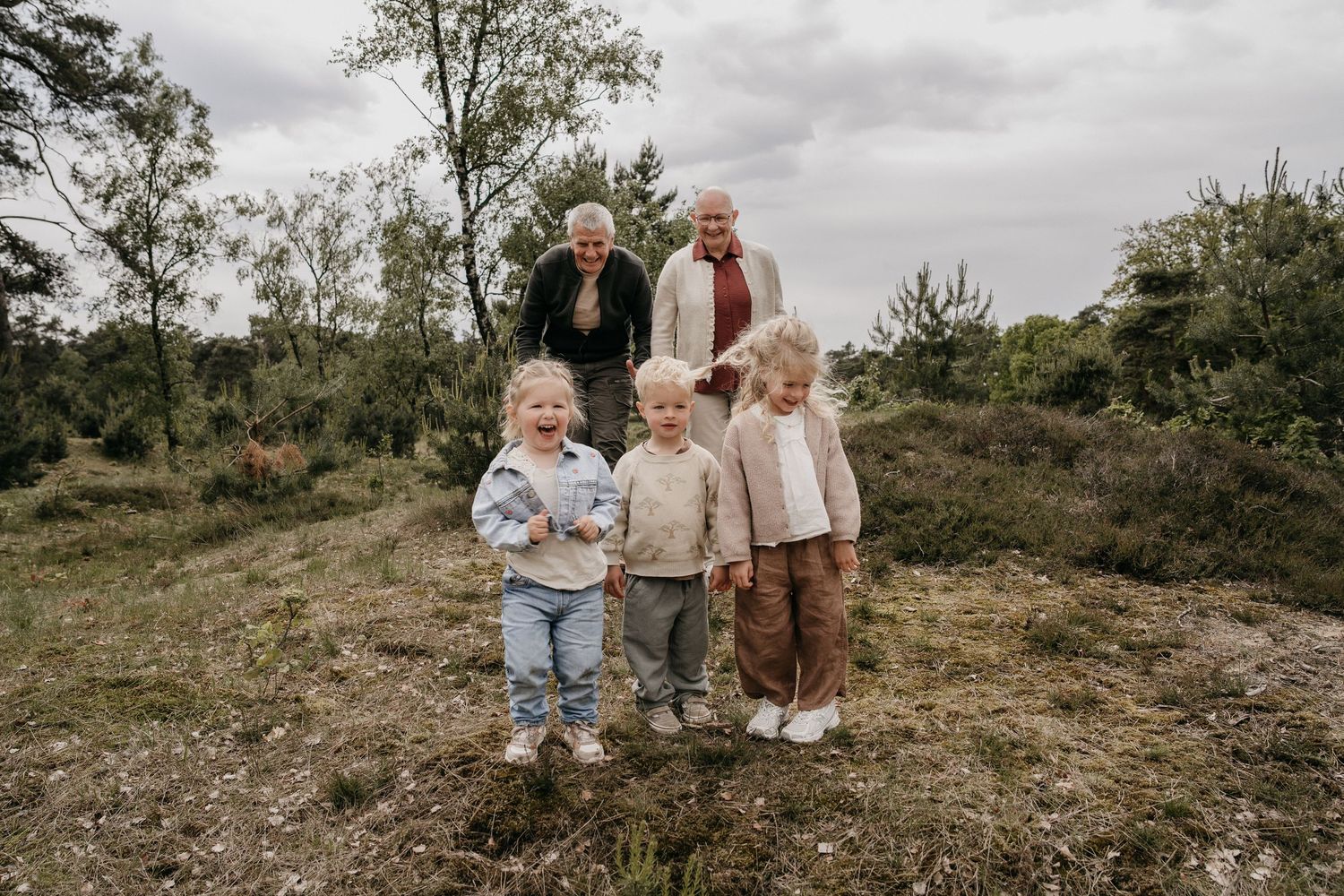 Grandparents smiling with three young toddlers outdoors on a grassy hill surrounded by trees on a cloudy day.