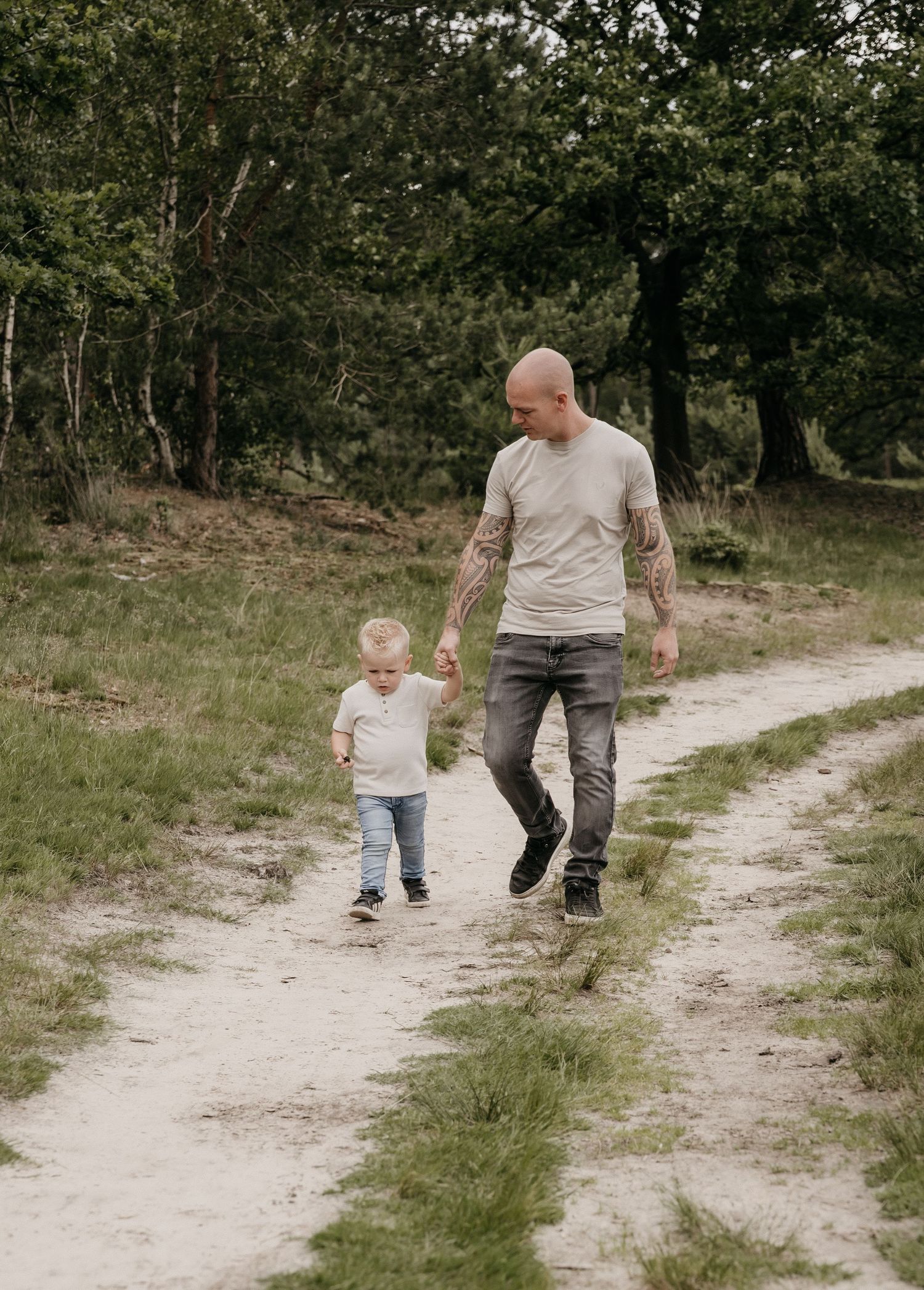A tattooed bald man walks with a toddler on a sandy forest path surrounded by green trees.