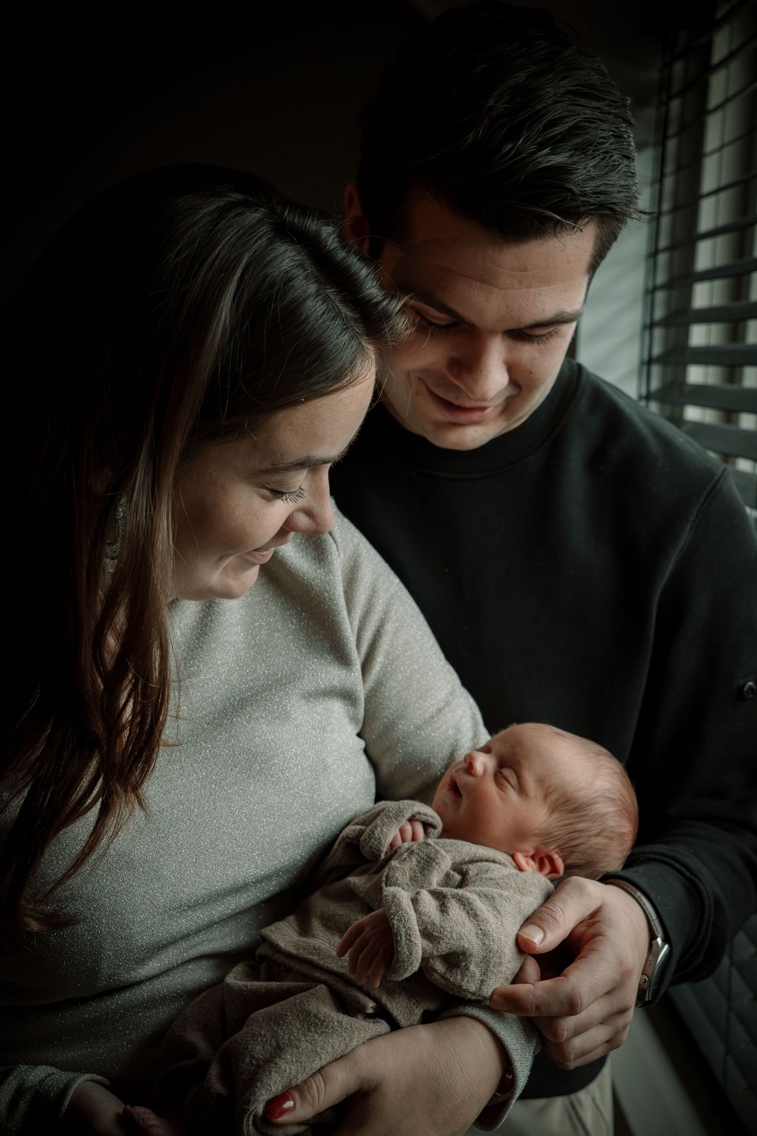 A couple shares an intimate moment with their newborn baby by a window in moody lighting.