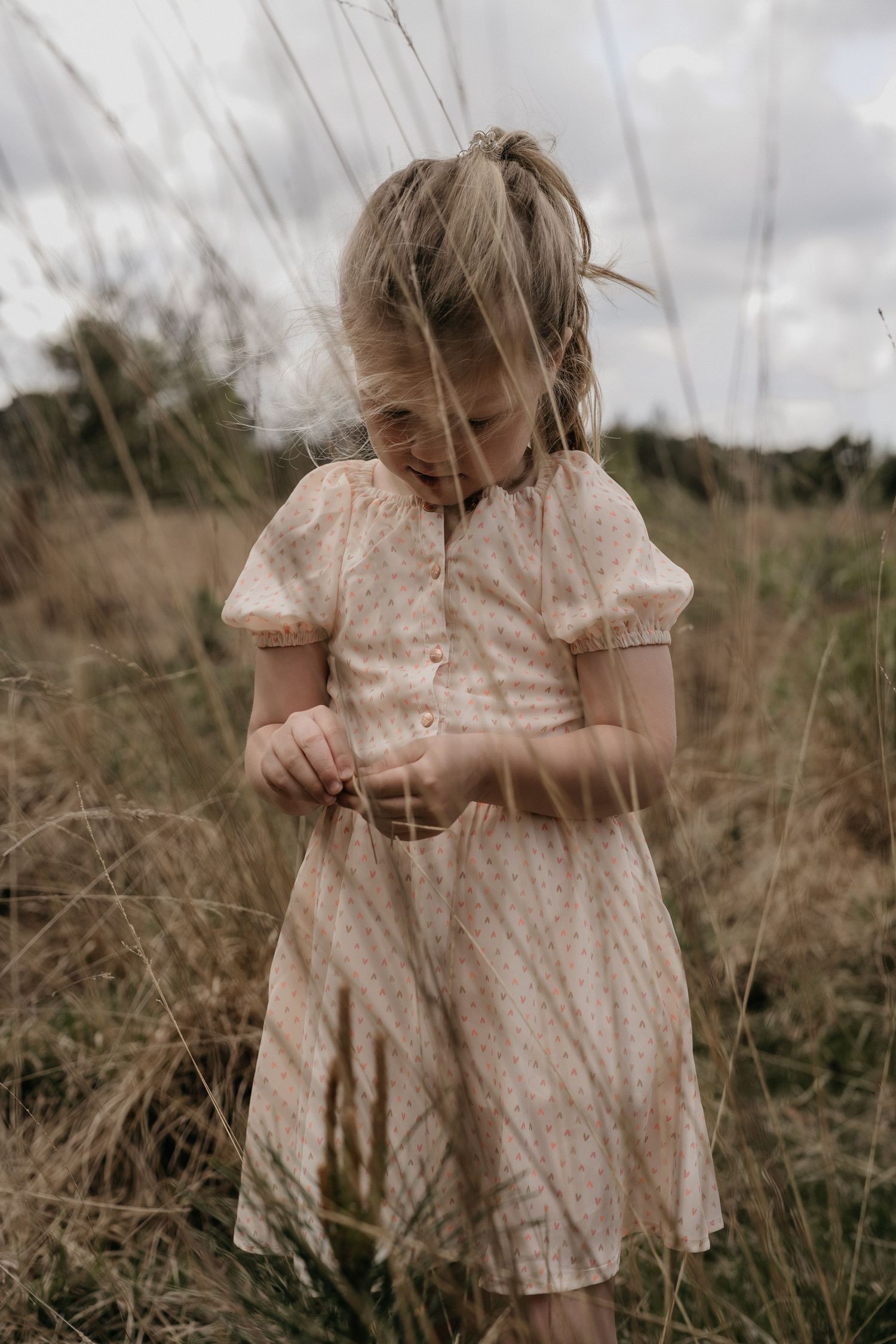 Young girl in floral dress standing in tall grass field on cloudy day.