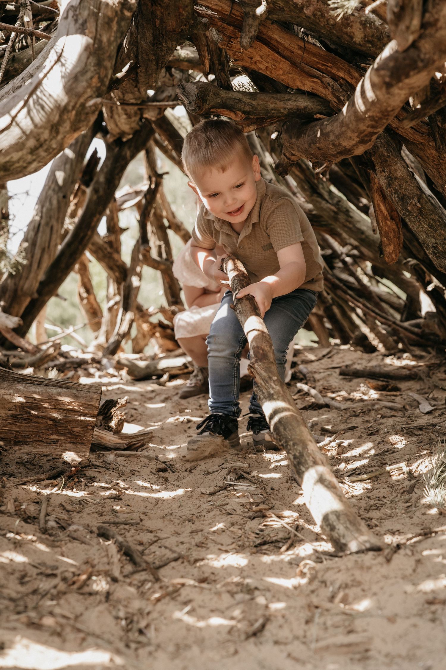 Young boy playing with sticks inside a rustic wooden fort structure outdoors.