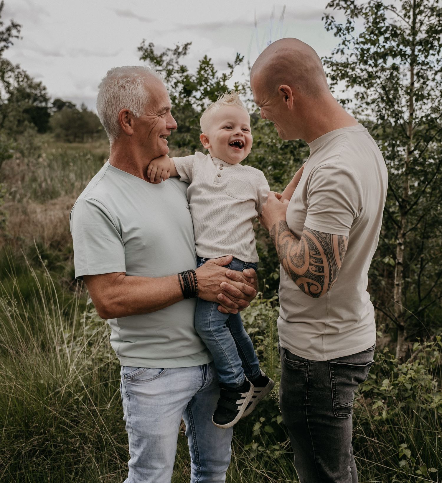 Two men and a laughing toddler share a joyful moment outdoors surrounded by lush greenery.