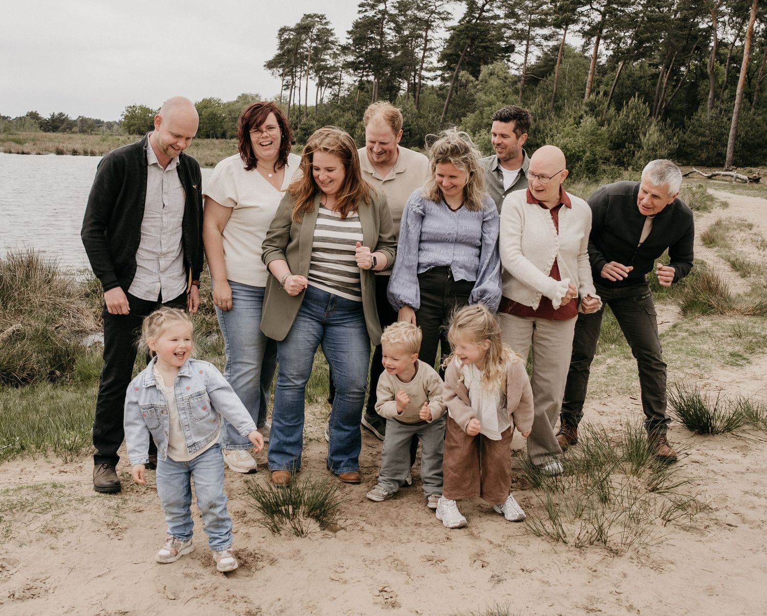 Multi-generational family laughing and posing together on a sandy beach path surrounded by trees and a lake.