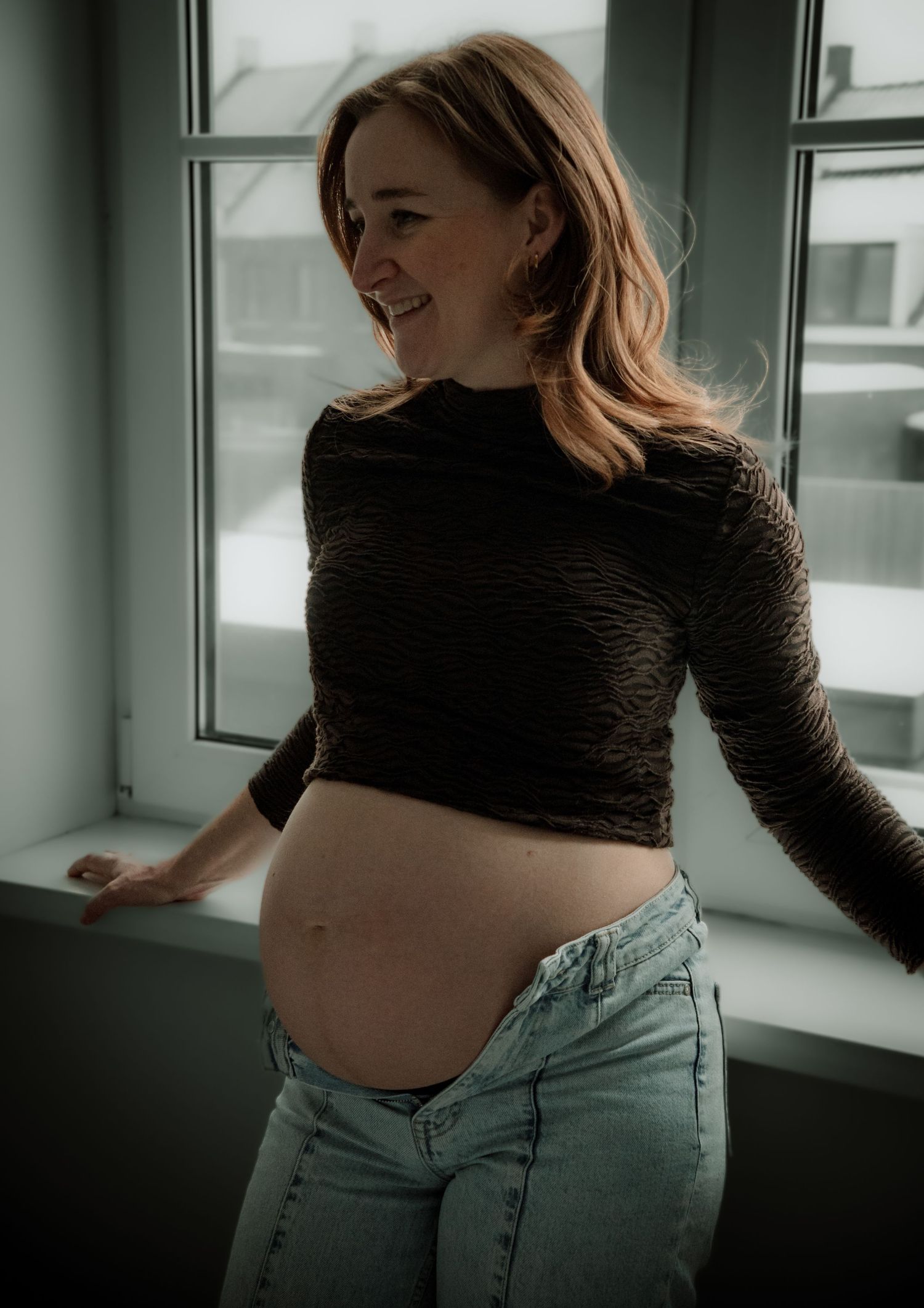 Smiling pregnant woman in crop top and jeans posing by window showing baby bump.