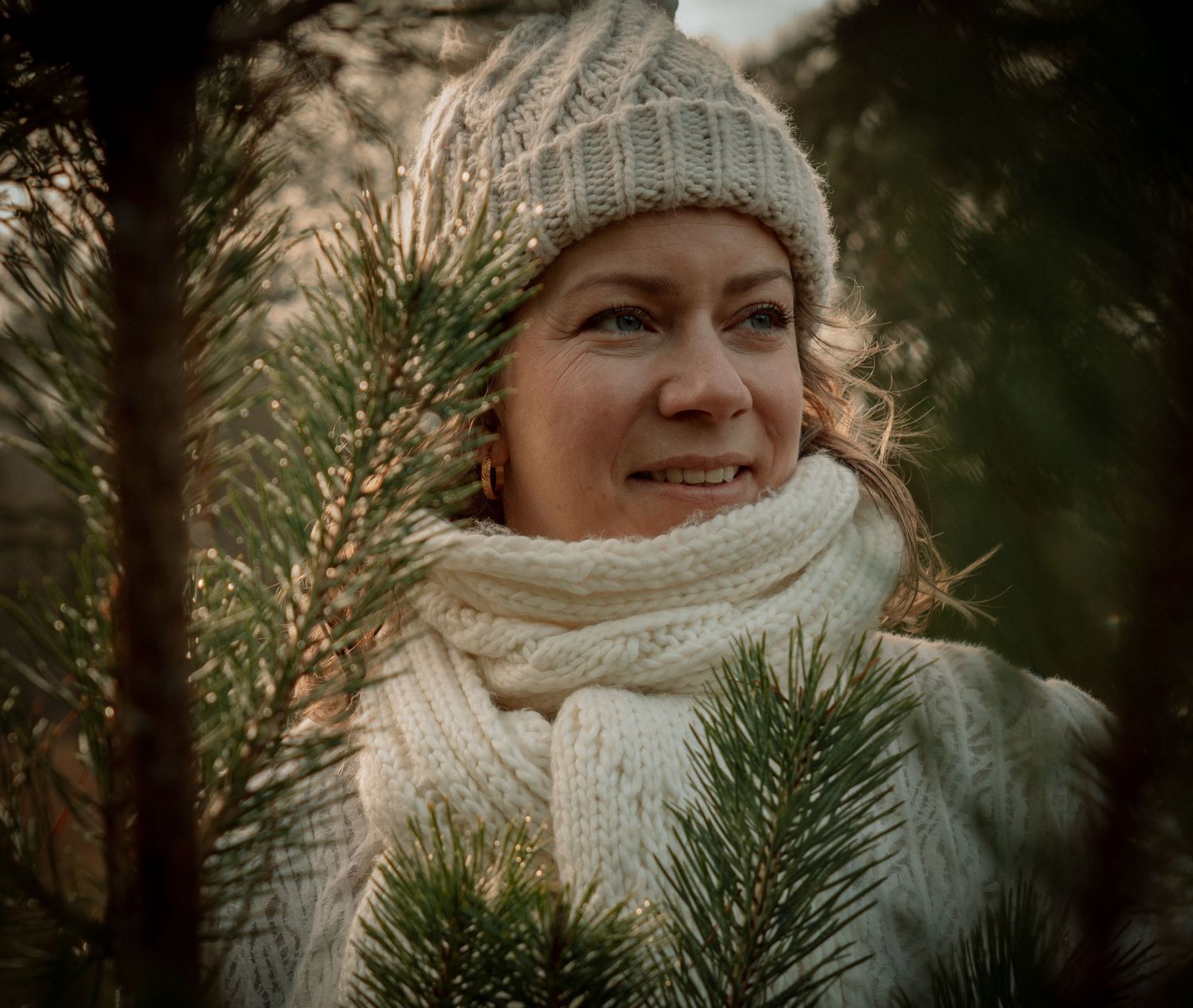 A person wearing a cozy gray knit hat and cream scarf stands among evergreen branches on a winter day.