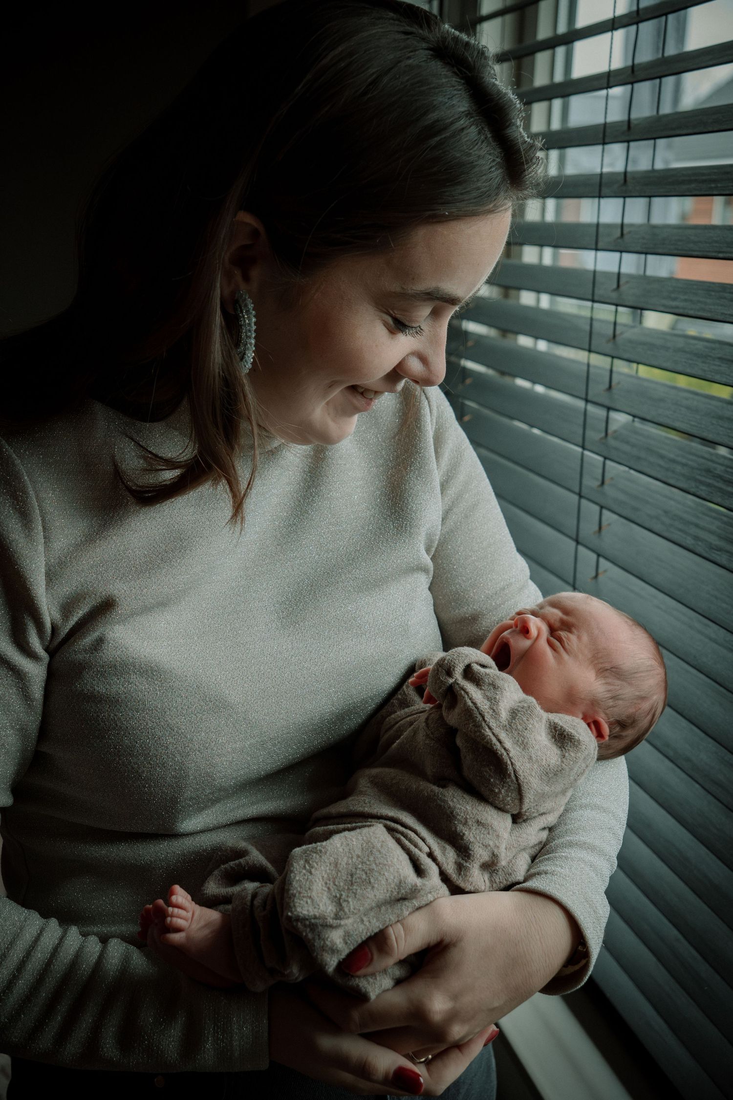 A tender moment by a window as a parent cradles and gazes lovingly at their sleeping newborn baby in soft lighting.