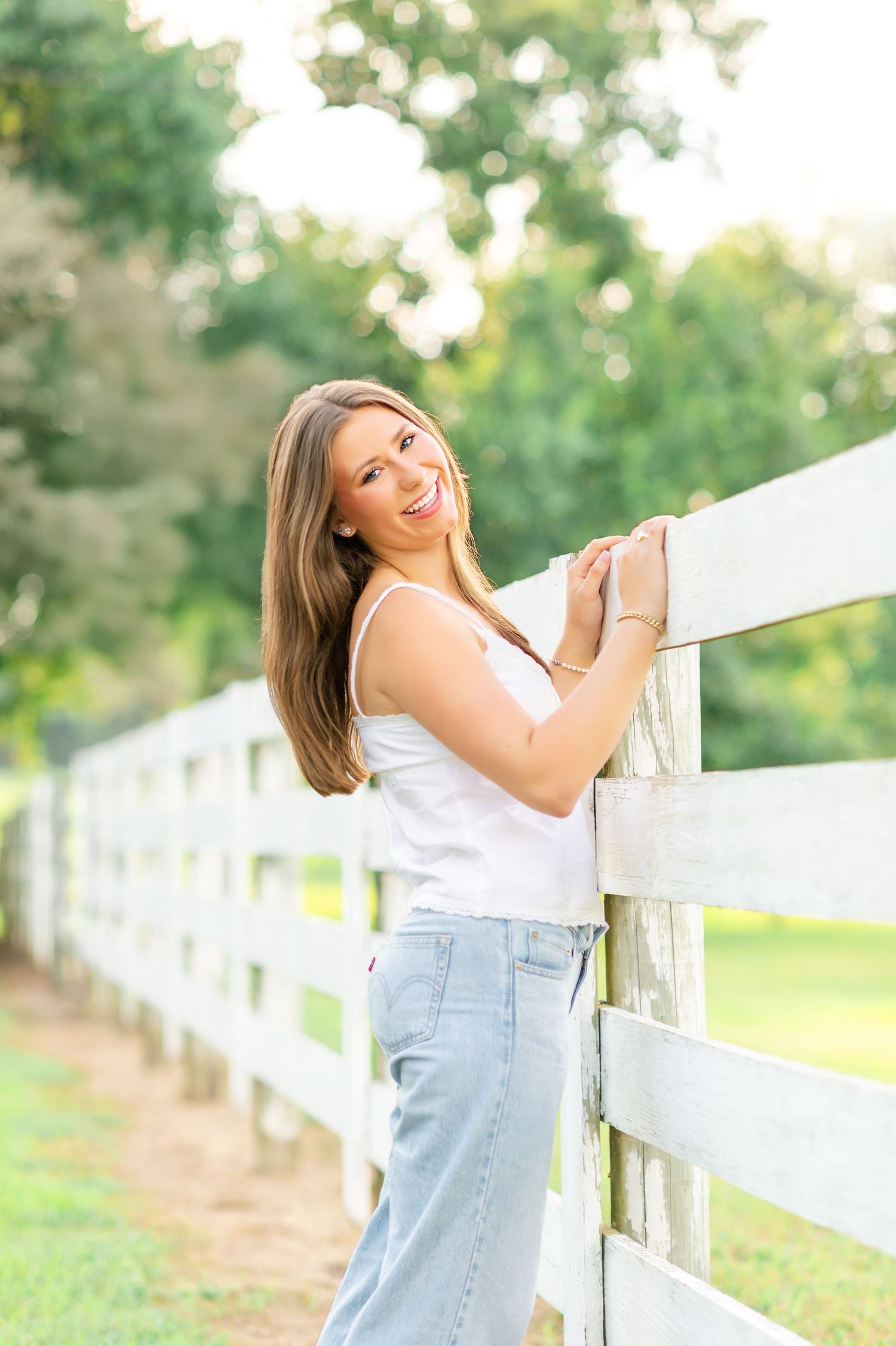 Smiling brunette teen leans on white wooden fence in white top and denim skirt at golden hour.