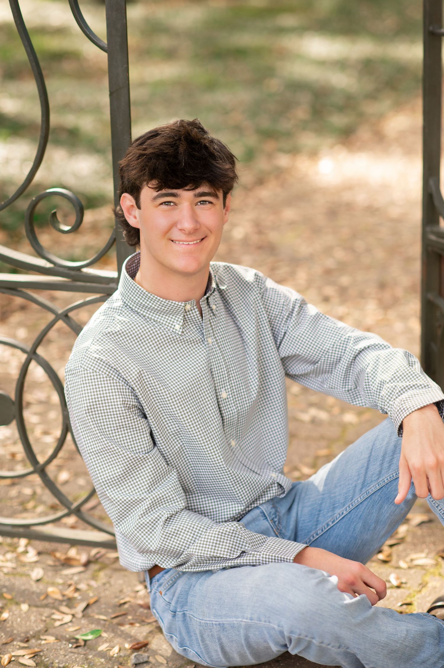 Smiling teenage boy in checkered shirt and jeans sitting cross-legged outdoors near decorative iron gate.