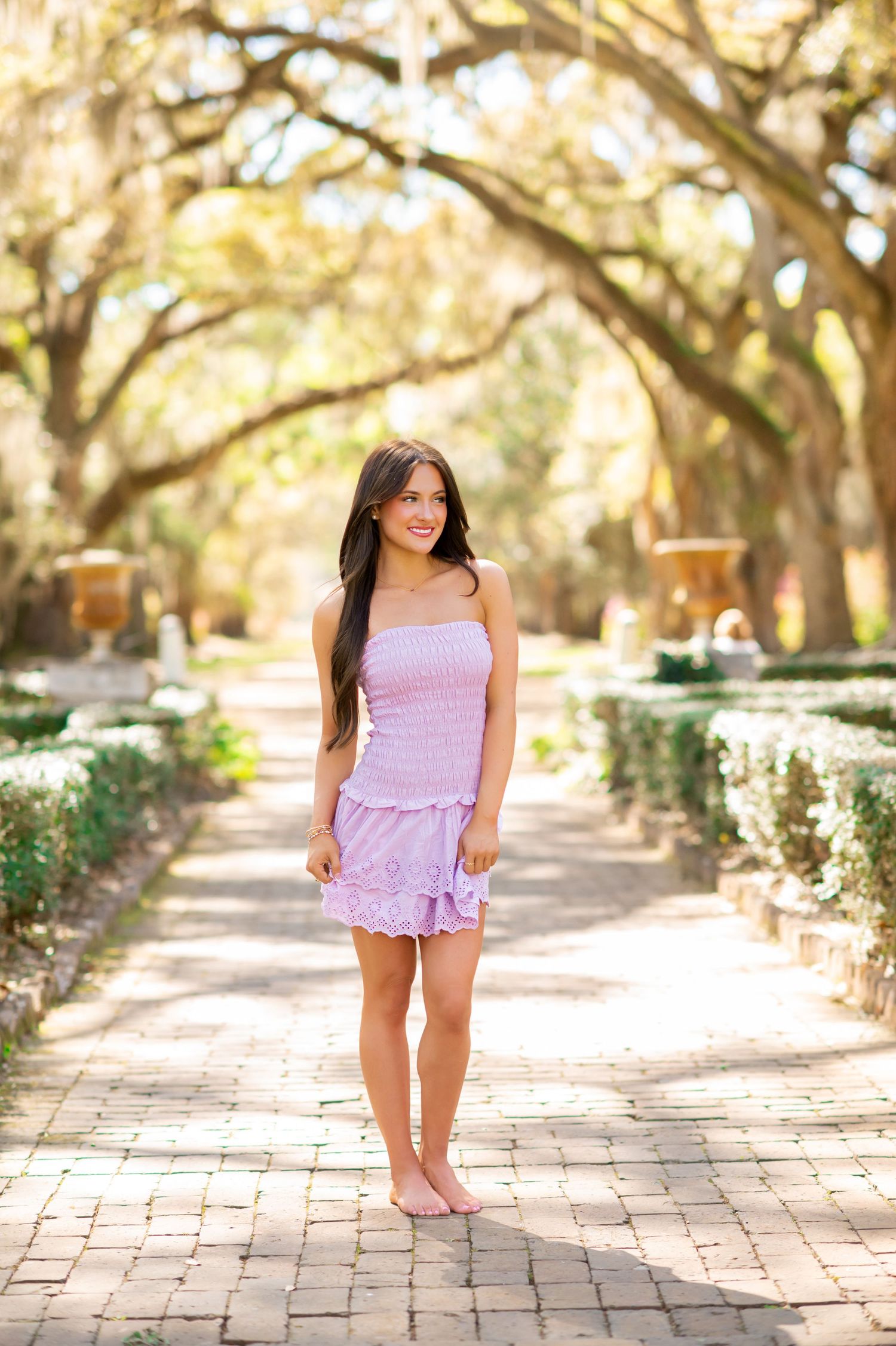 Young woman in lavender smocked mini dress standing on brick path lined with oak trees in a sunny garden.