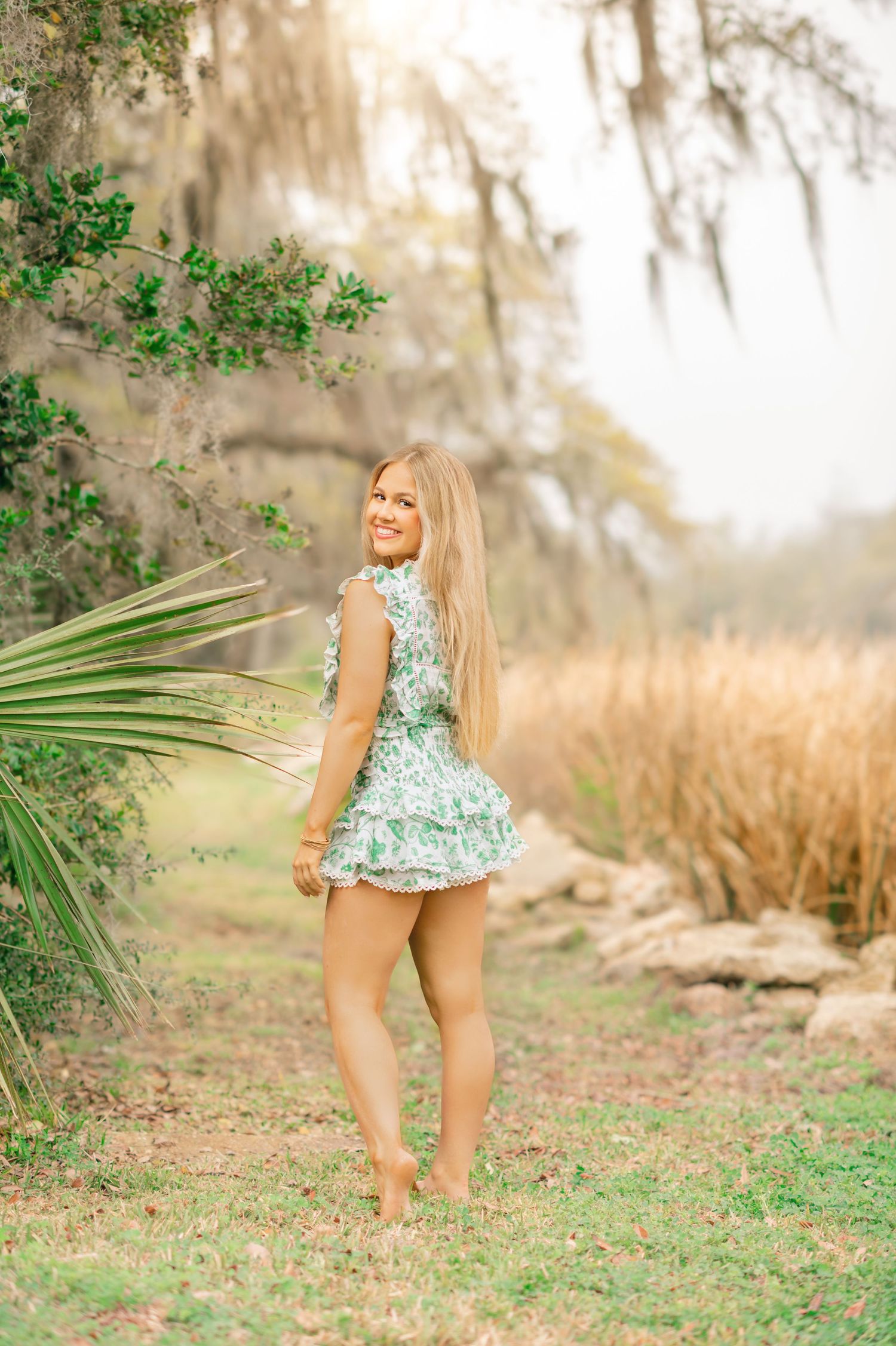 Blonde woman in green floral romper smiles over shoulder in misty outdoor setting with palm fronds.