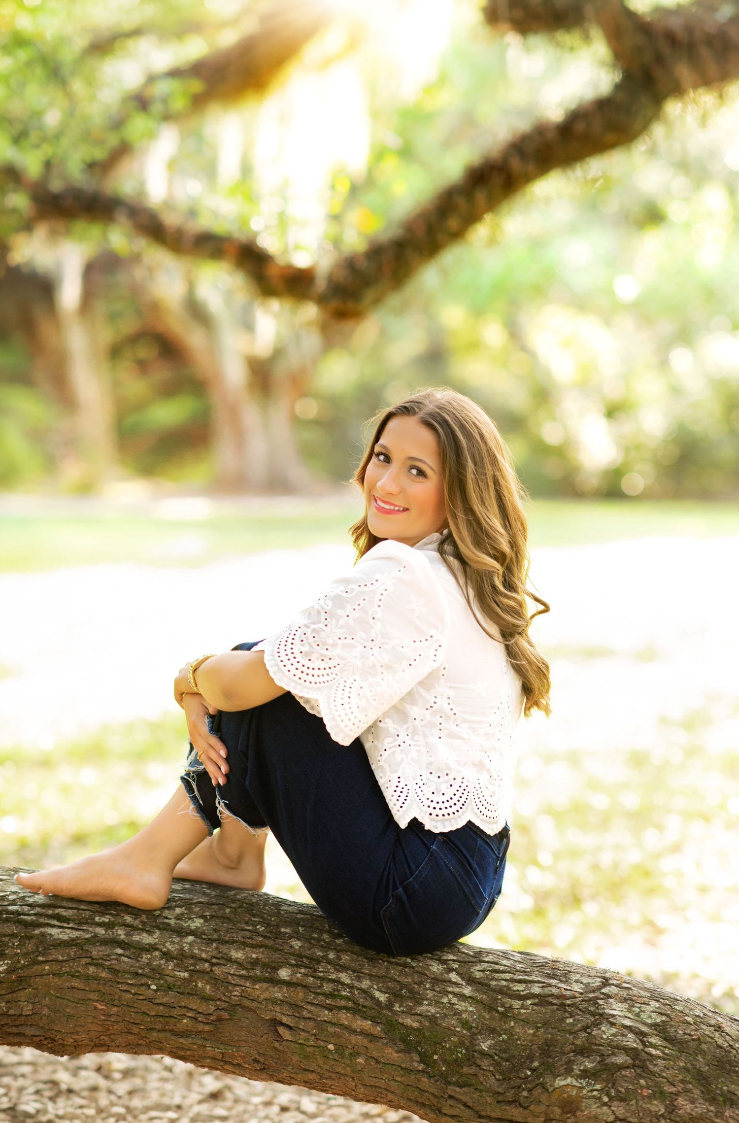 Young woman in white lace top and jeans sitting on a tree log in a sunlit park, smiling over her shoulder.