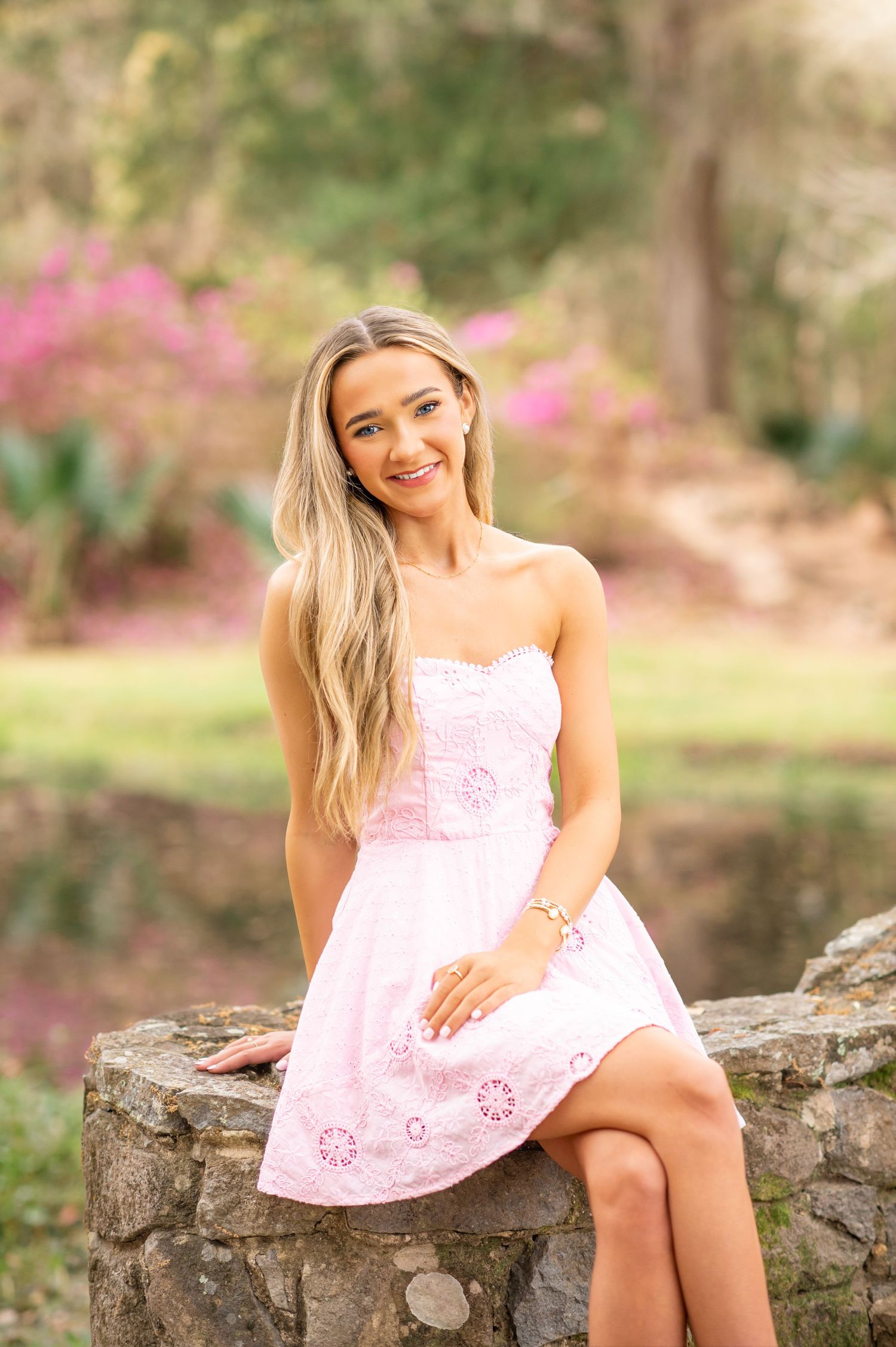 Young woman in pink strapless dress sitting on stone wall in garden with blooming flowers.