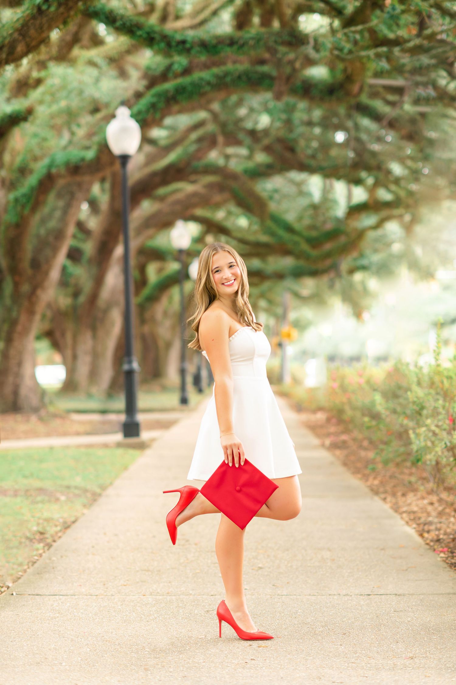 Young woman in white dress and red heels holding graduation cap on tree-lined sidewalk path.