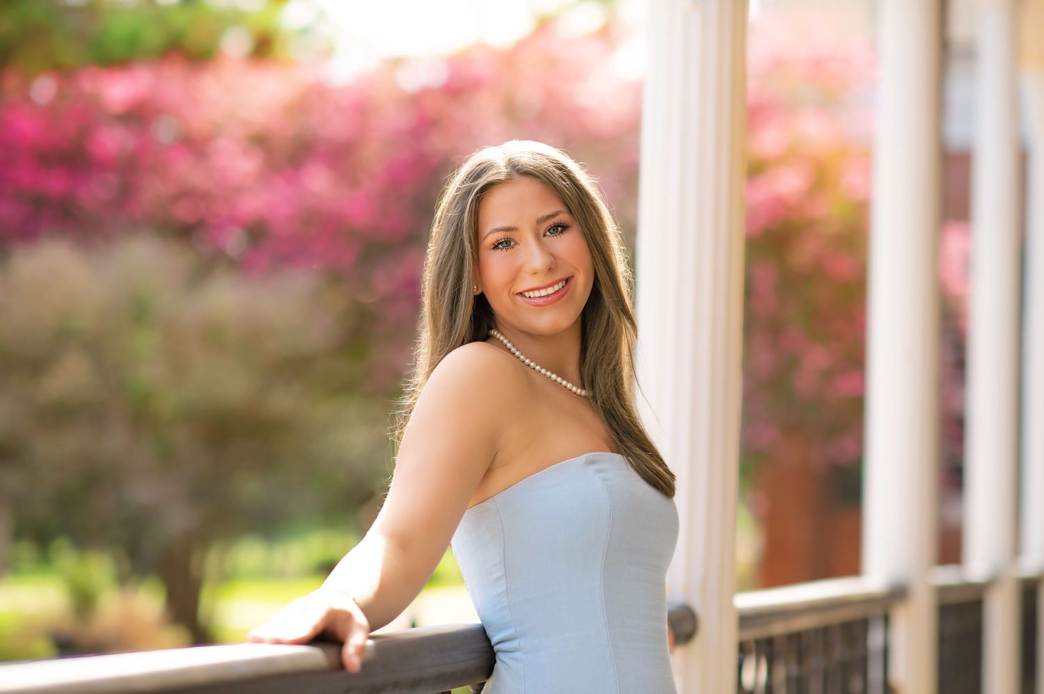 Young woman in light blue strapless dress smiling on a balcony with pink flowering trees in background.