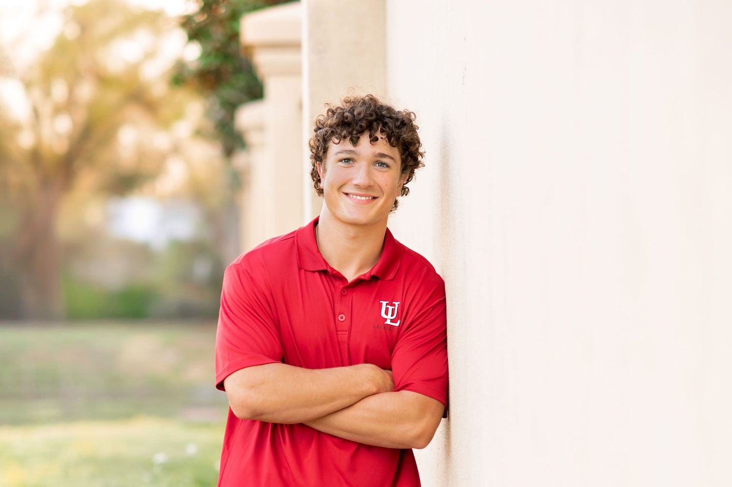 Young man in red University of Utah polo shirt leaning against white column with arms crossed, smiling outdoors.