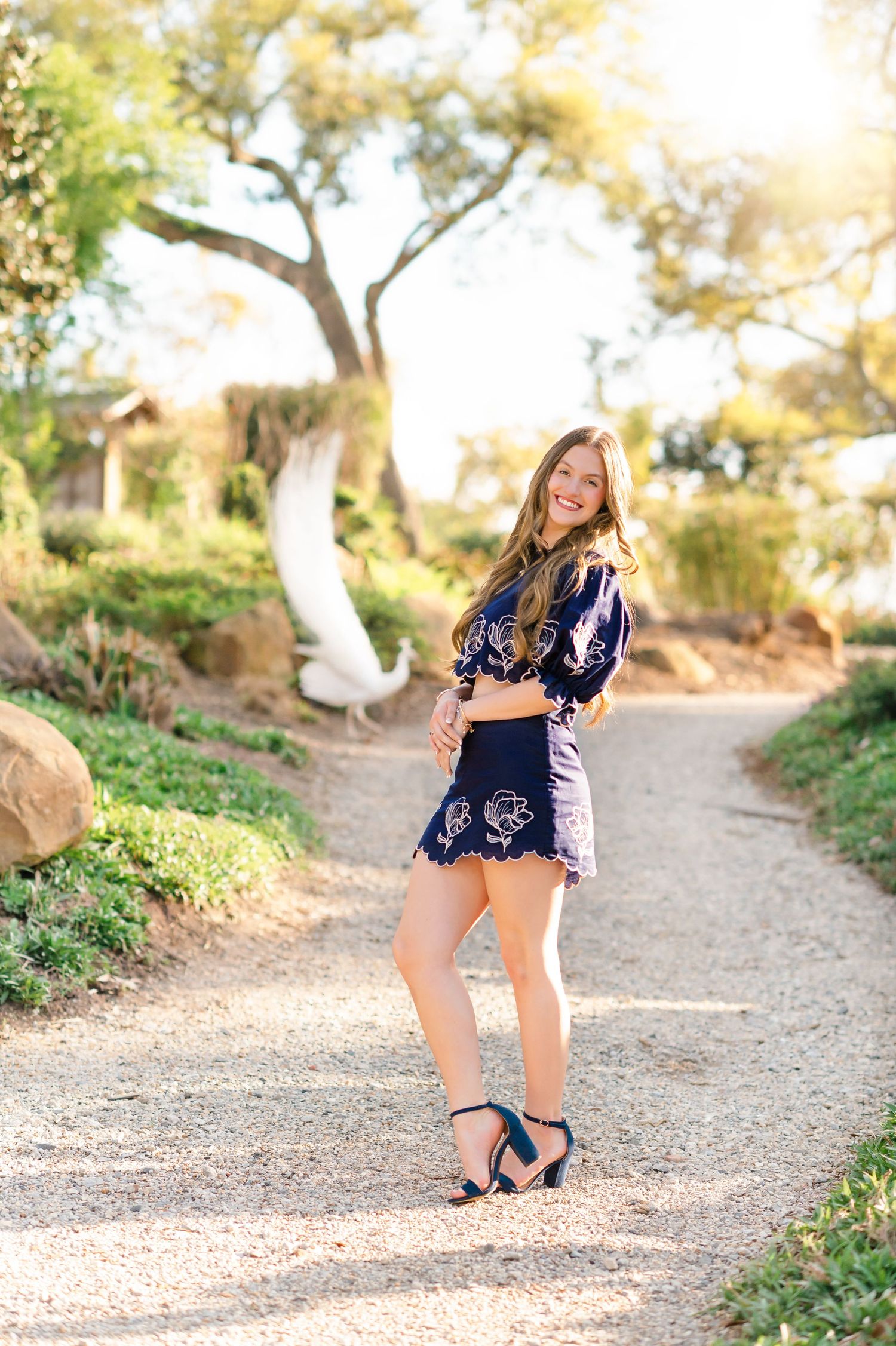 Young woman in navy floral crop top and skirt with teal heels posing on a sunlit garden path.