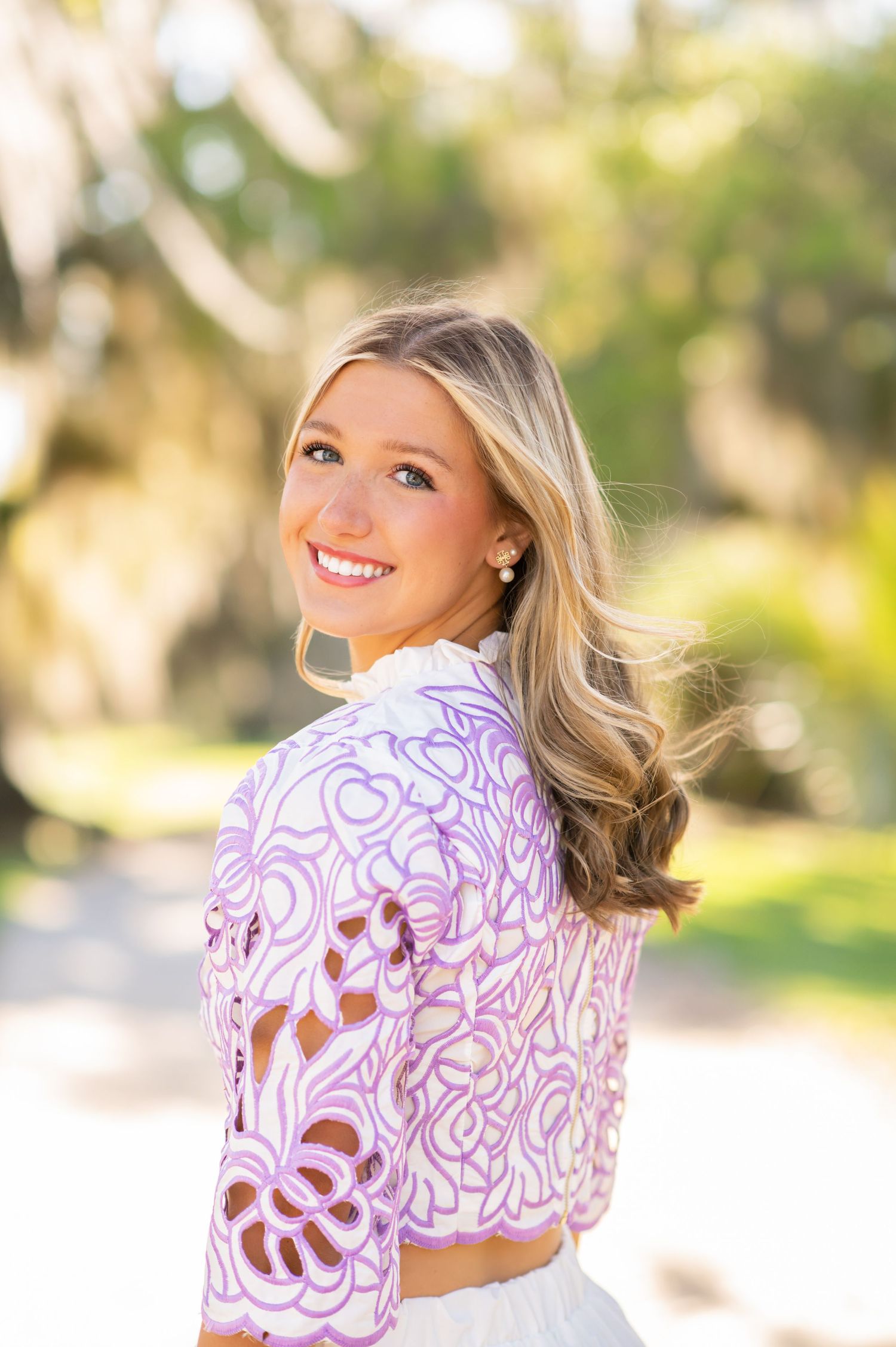 Young blonde woman smiling over shoulder in purple floral lace crop top outdoors with green trees in background.