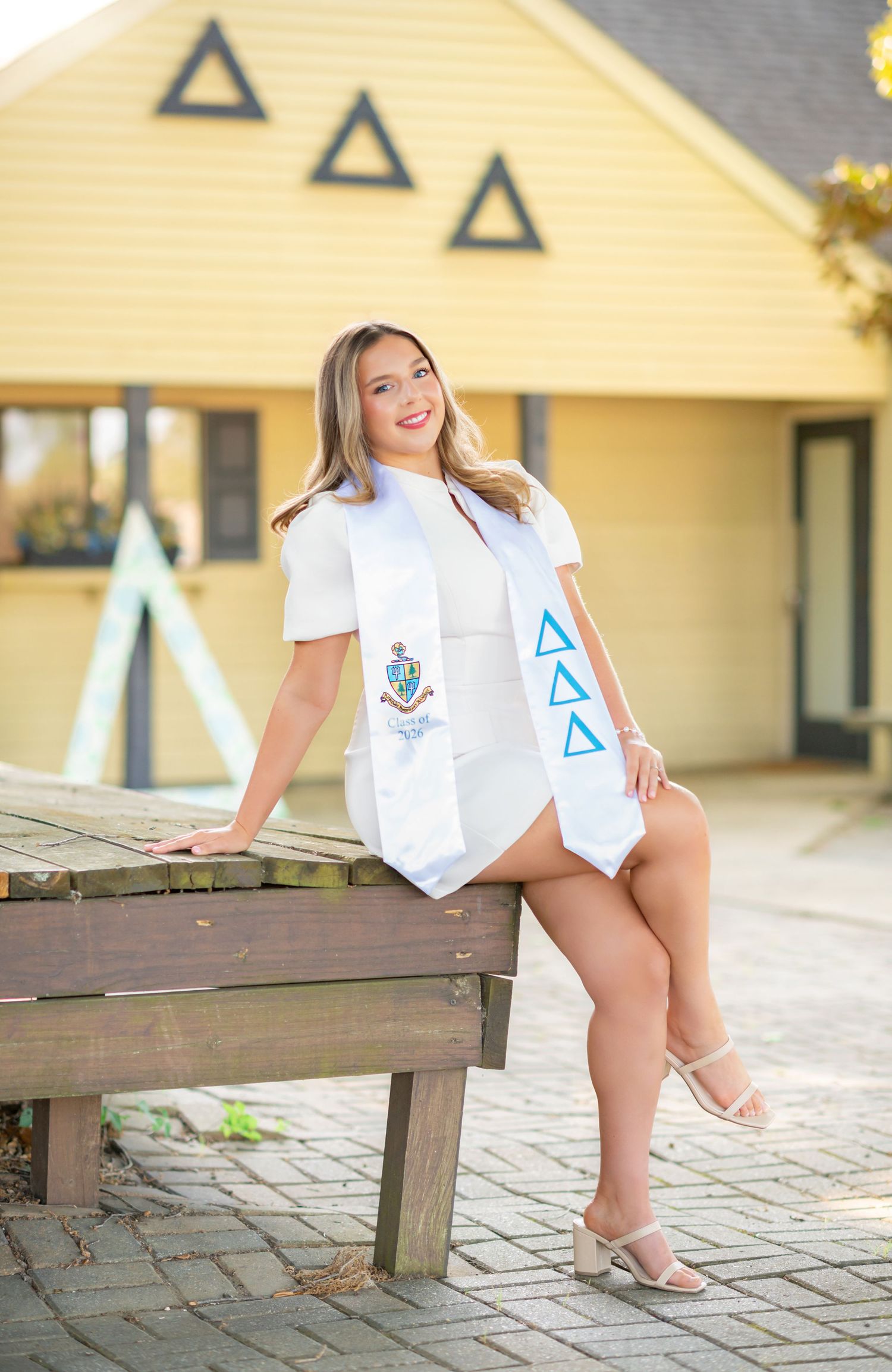 Smiling graduate poses in white dress with Delta Delta Delta sorority sash on wooden bench outside yellow house.