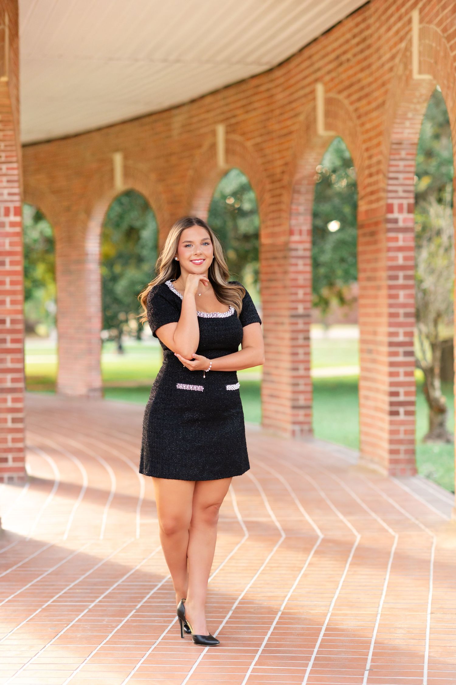 Young woman in black mini dress with pink accents posing under brick arches on a university campus.