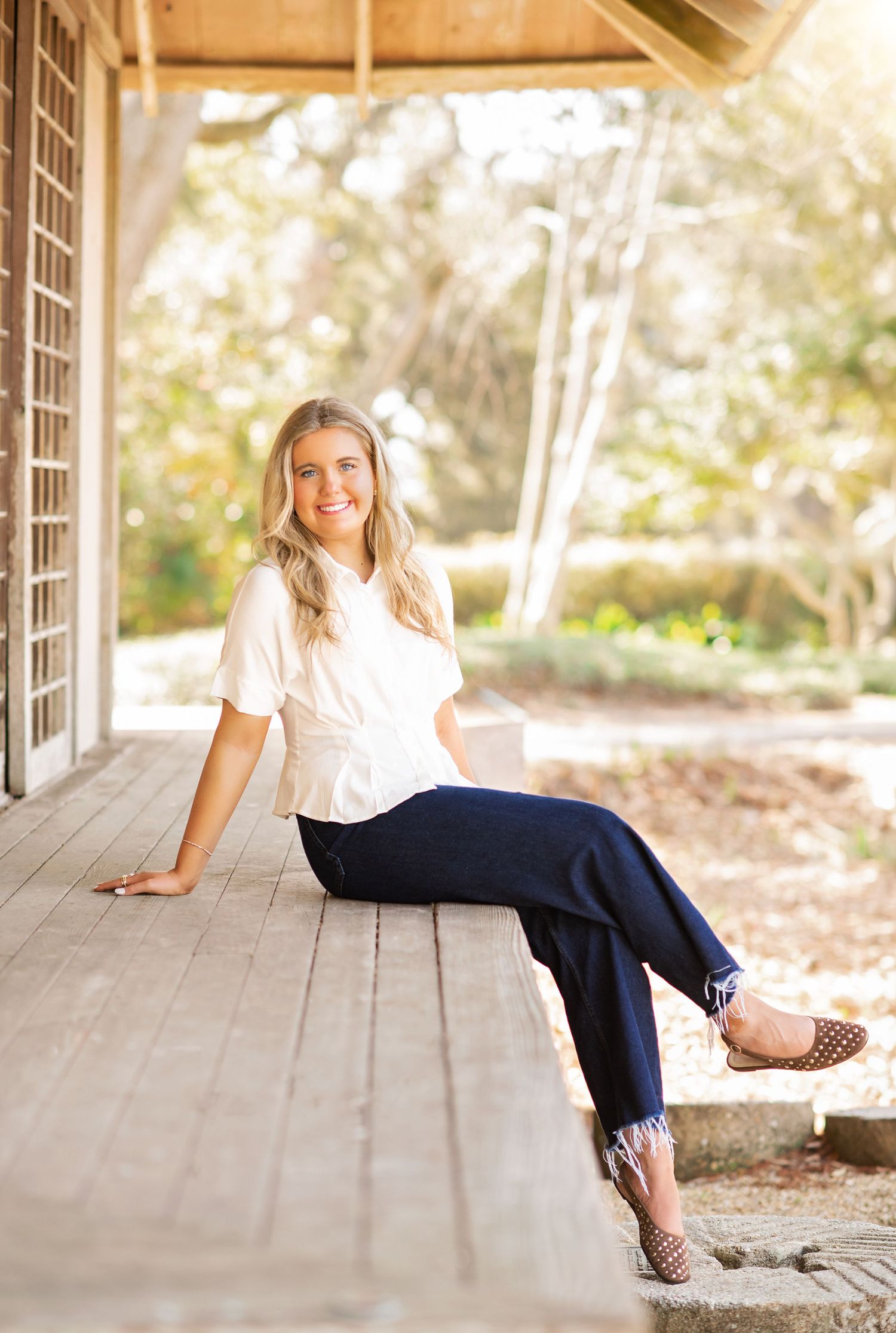 Young blonde woman smiling, seated on wooden porch steps in white top and navy pants with leopard flats.