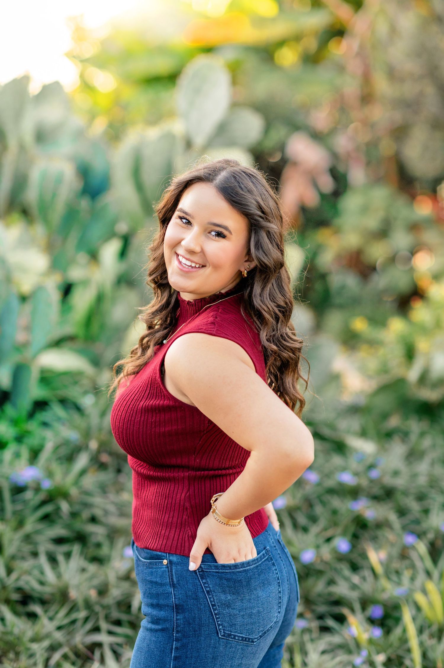 Young woman in red top and jeans smiling outdoors near cacti during golden hour senior portrait.