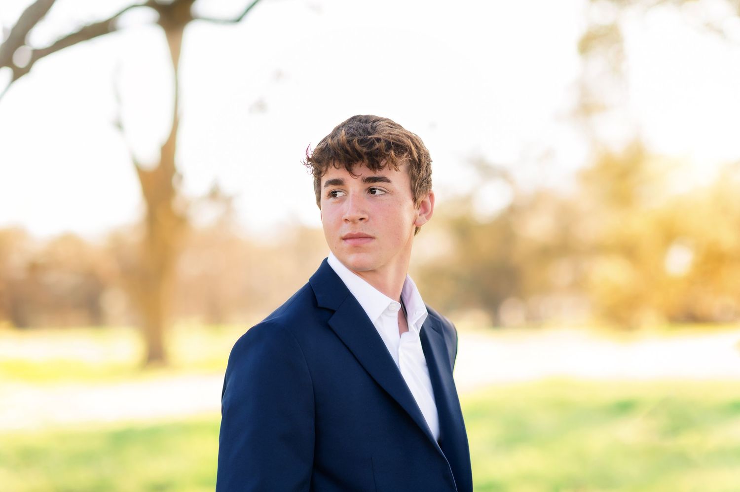 Young man in navy blue suit posing outdoors in a park during golden hour for senior portraits.