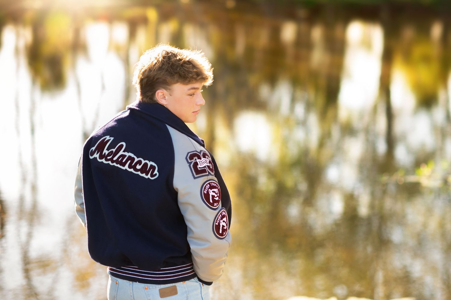 Teen boy in a varsity letterman jacket standing near a reflective pond in golden hour light.