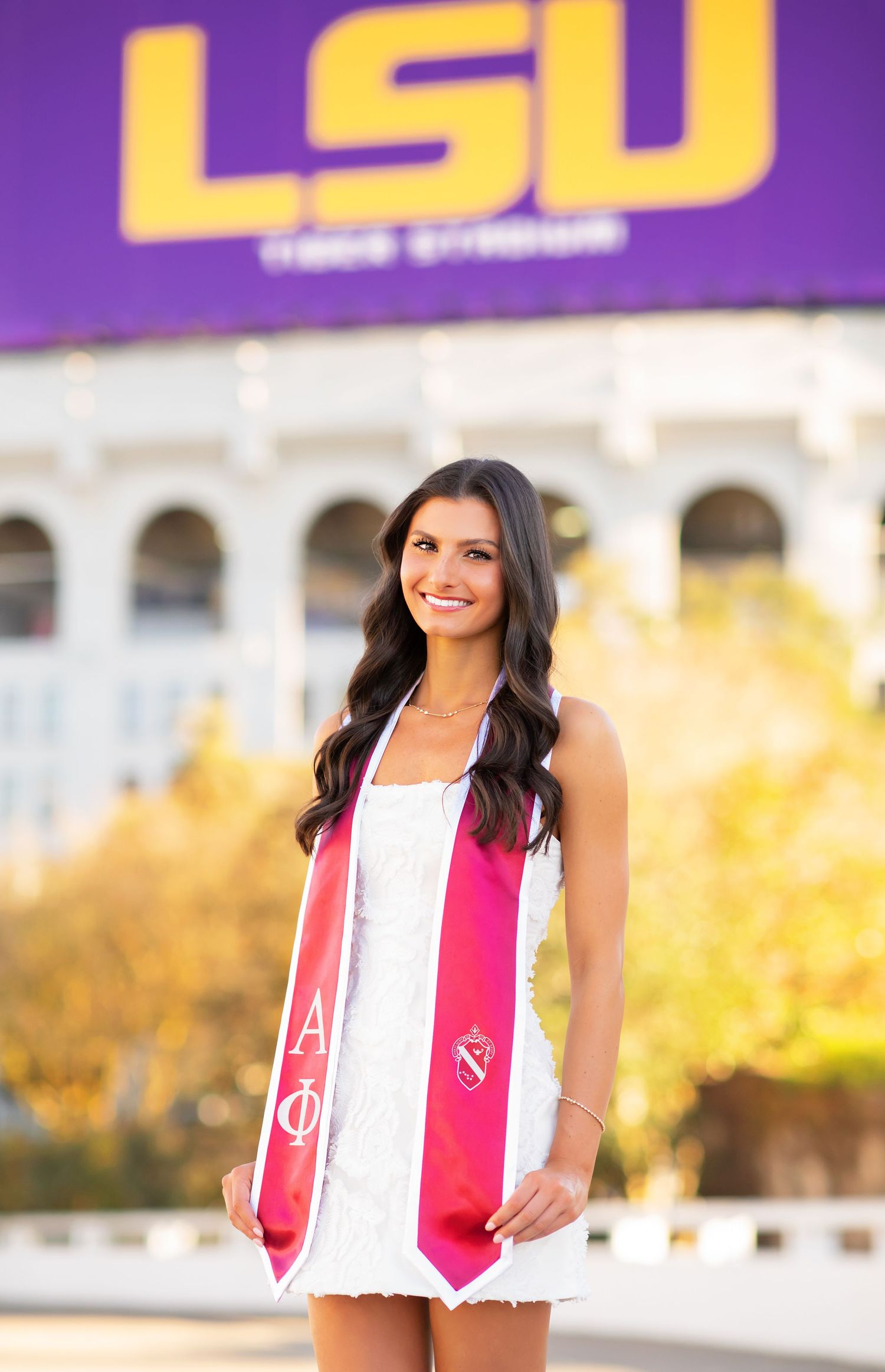 LSU graduate wearing Alpha Phi sash poses in white dress before Tiger Stadium.