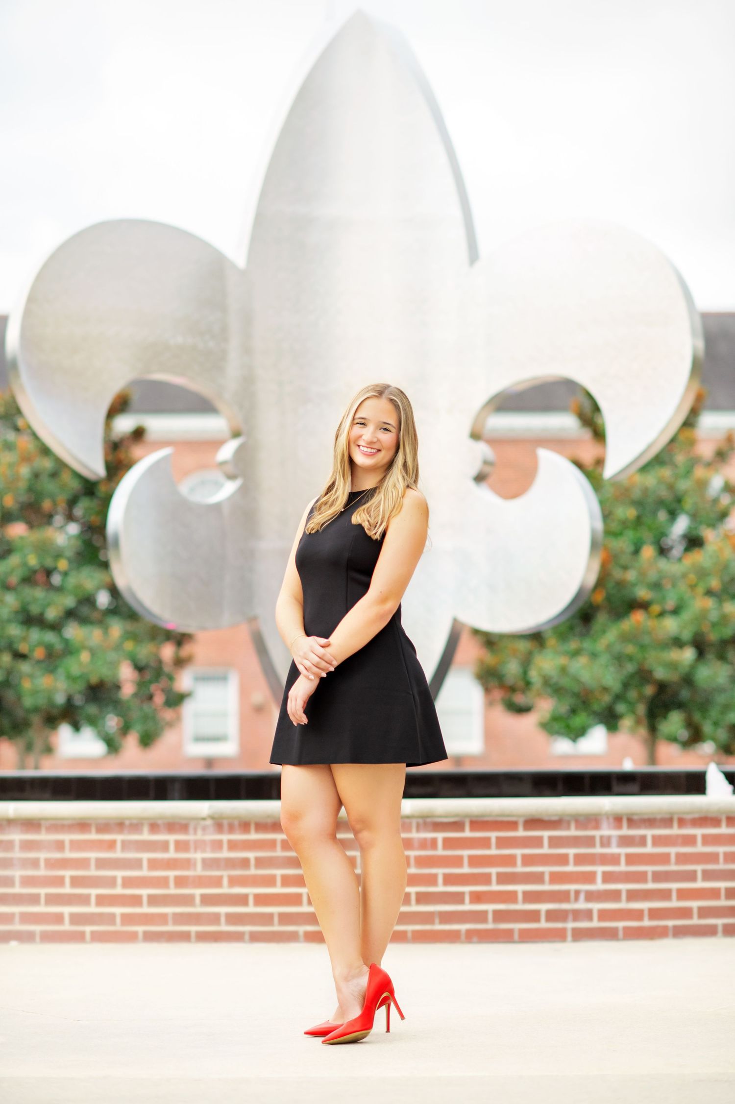 Young woman in black dress and red heels posing before a large fleur-de-lis sculpture outdoors.
