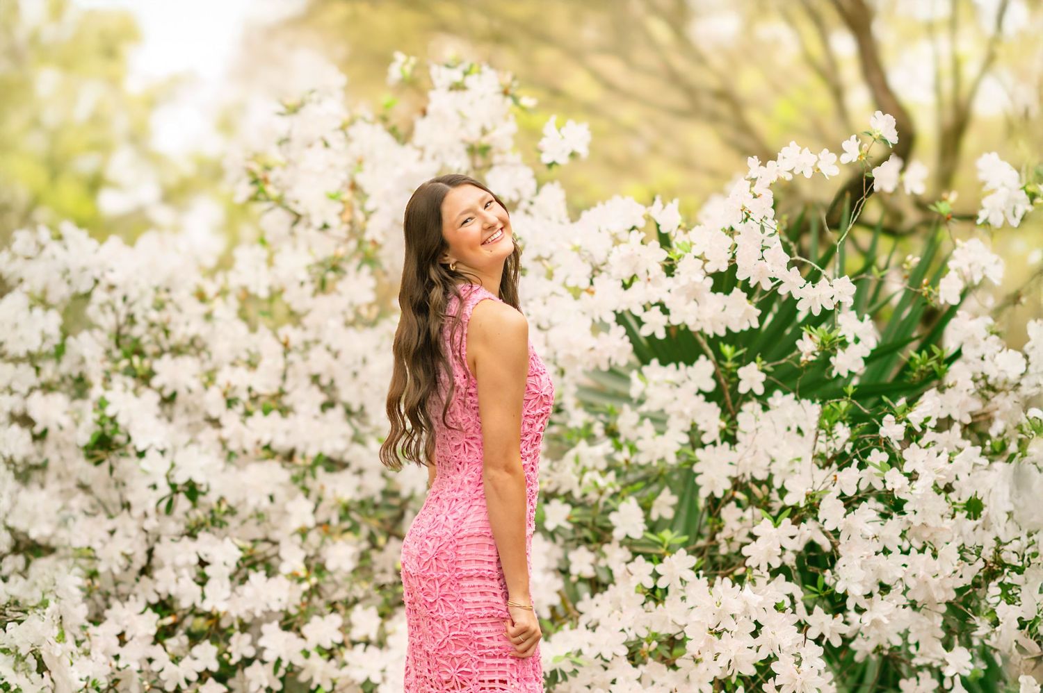 Young woman in pink dress smiles among white flowering bushes in a bright outdoor garden setting.