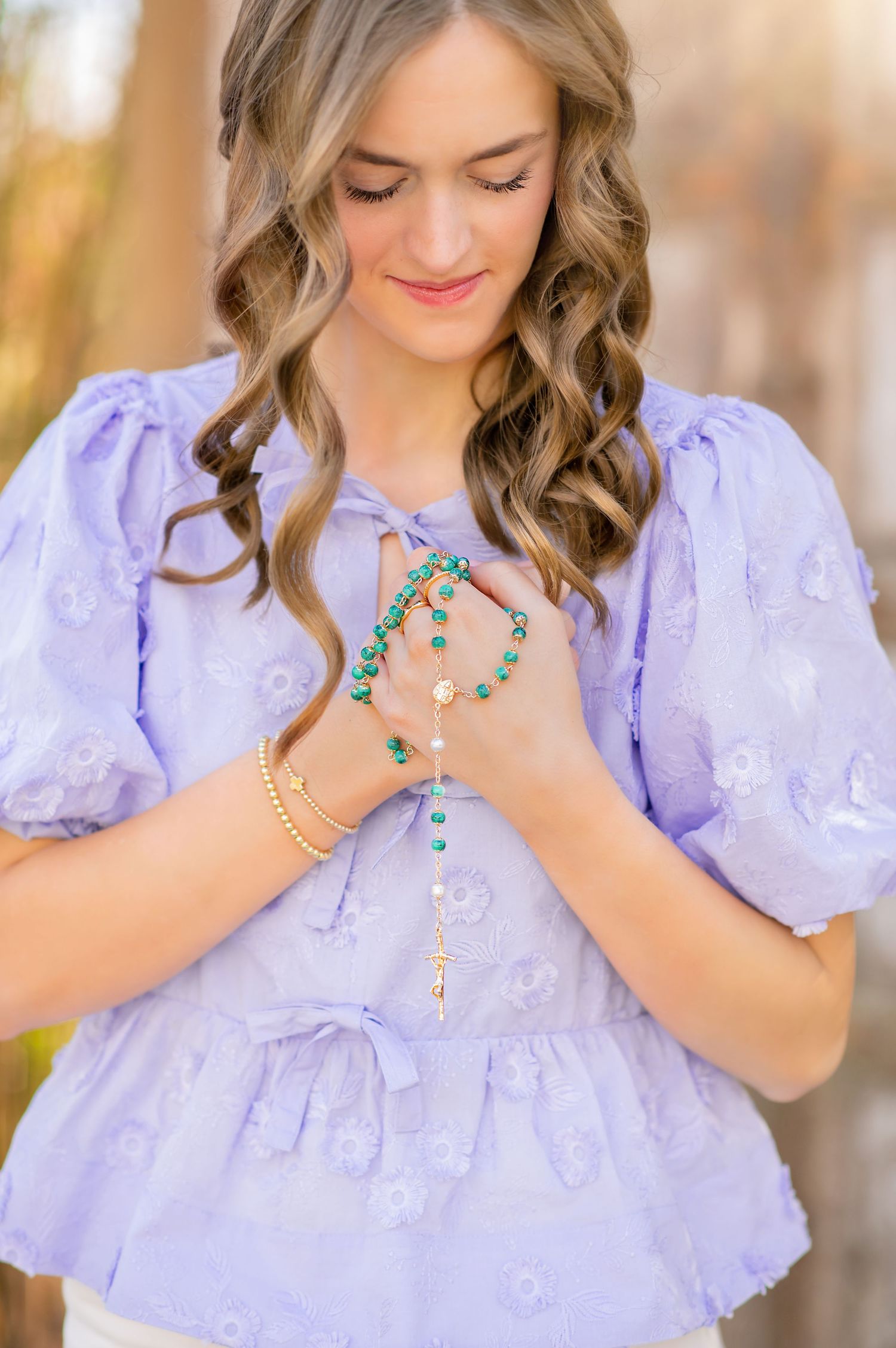 Young woman in lavender puff-sleeve top holding green rosary beads with hands clasped in prayer.