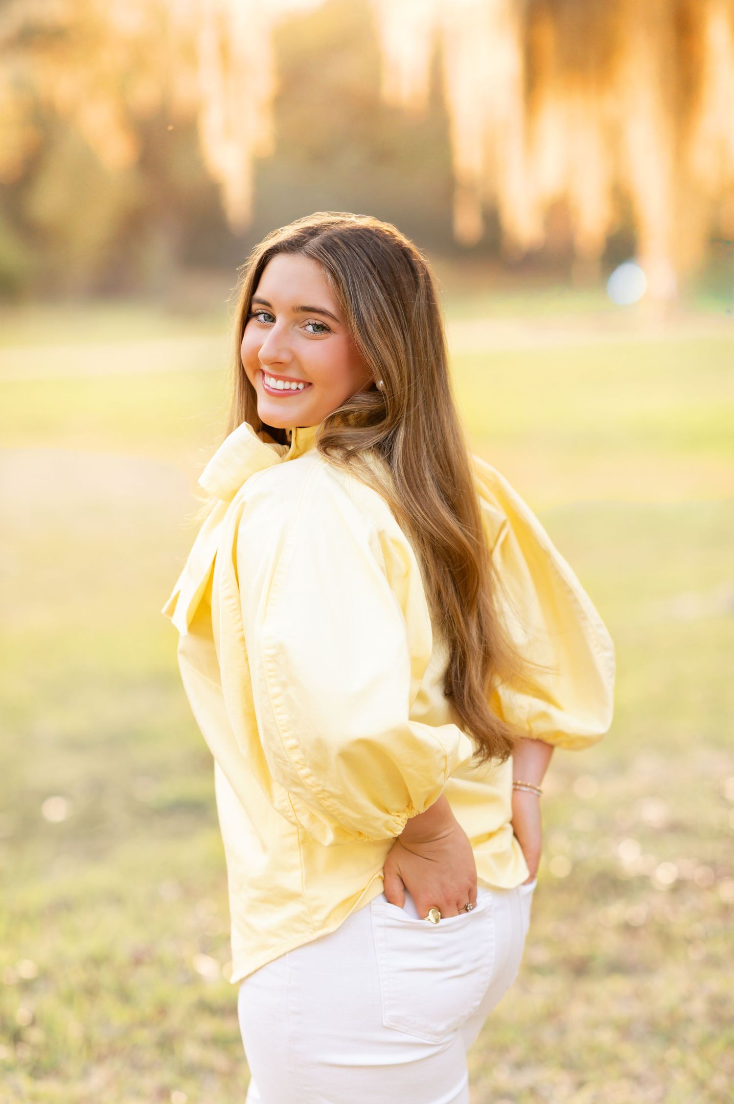 Young woman in yellow blouse and white pants smiling outdoors in golden sunlight.